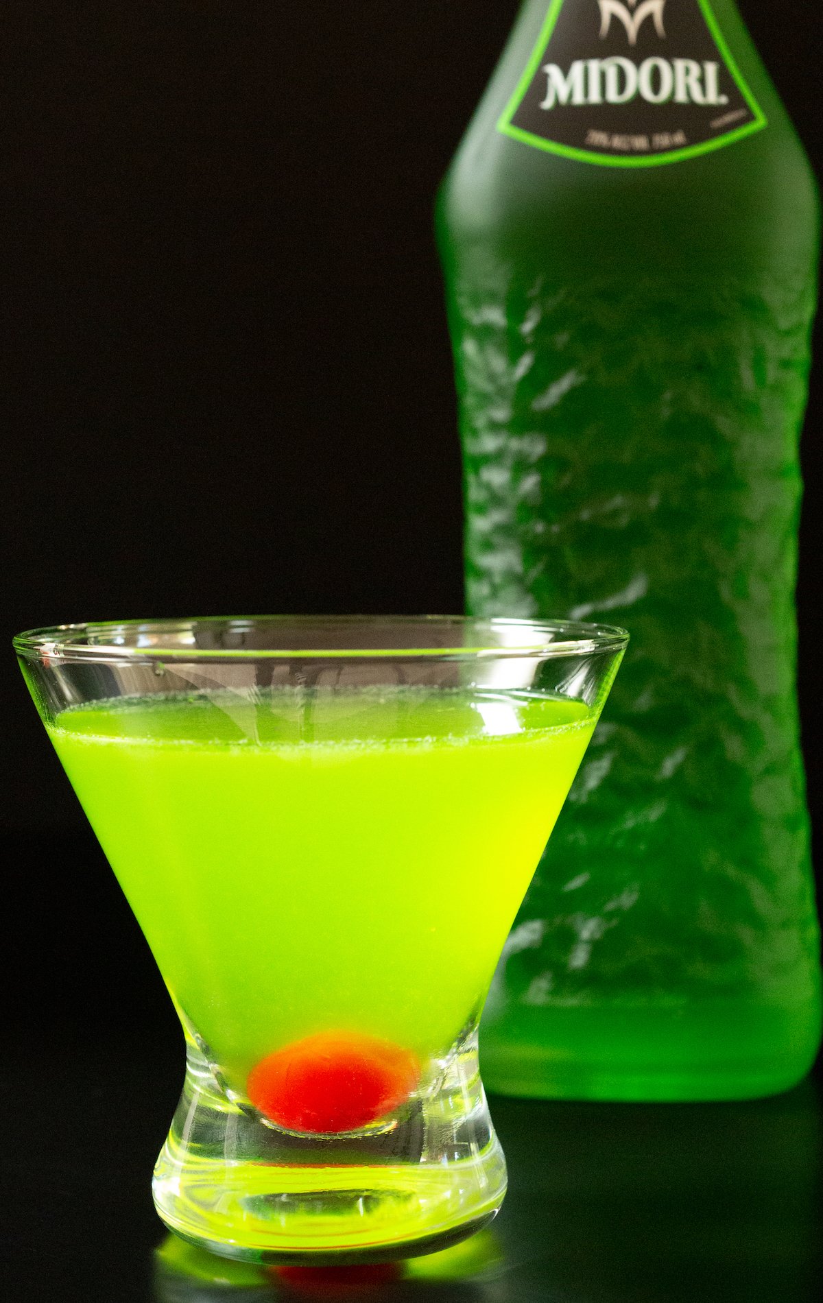 A stemless martini glass filled with a neon green Japanese slipper cocktails that has a maraschino cherry in the bottom. A bottle of Midori is out of focus in the background.