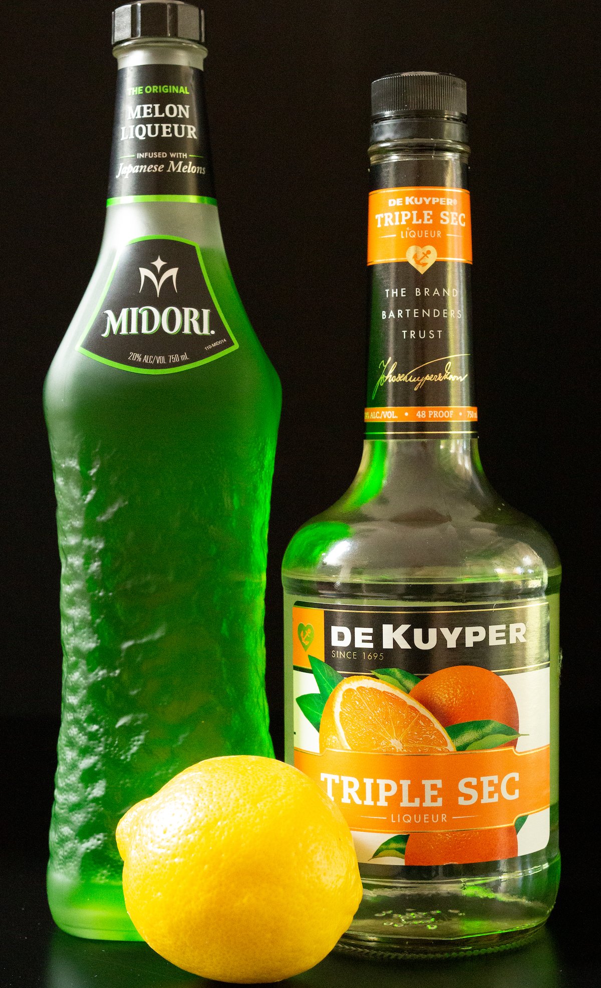 A bottle of Midori, triple sec, and lemon juice on a black background.