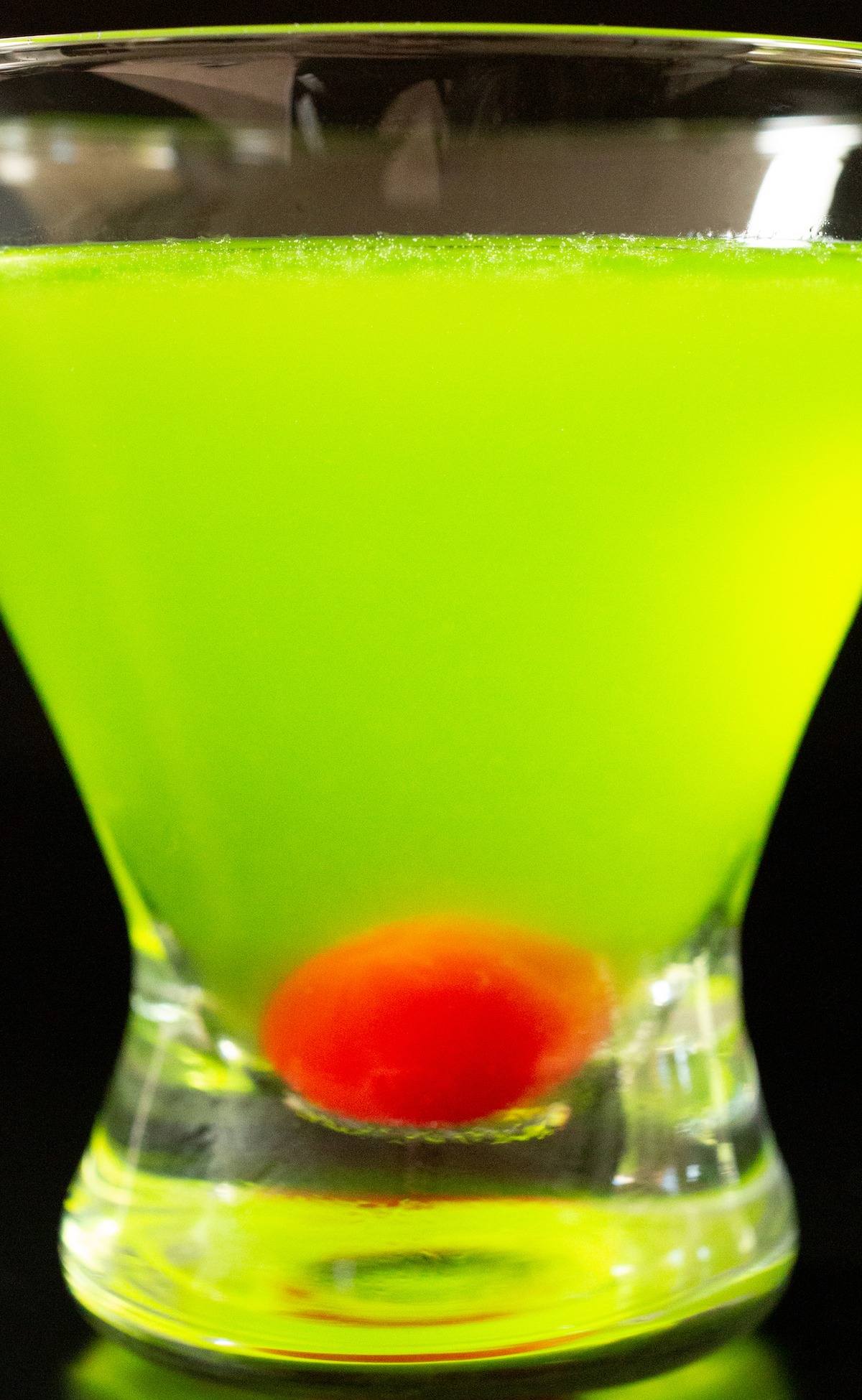 A stemless martini glass is filled with a neon green japanese slipper cocktail. There is a stemless maraschino cherry in the bottom of the glass.