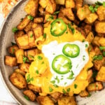 A serving bowl filled with chopped seasoned fiesta potatoes covered in nacho cheese and sour cream. Slices of jalapeno and chives on top.