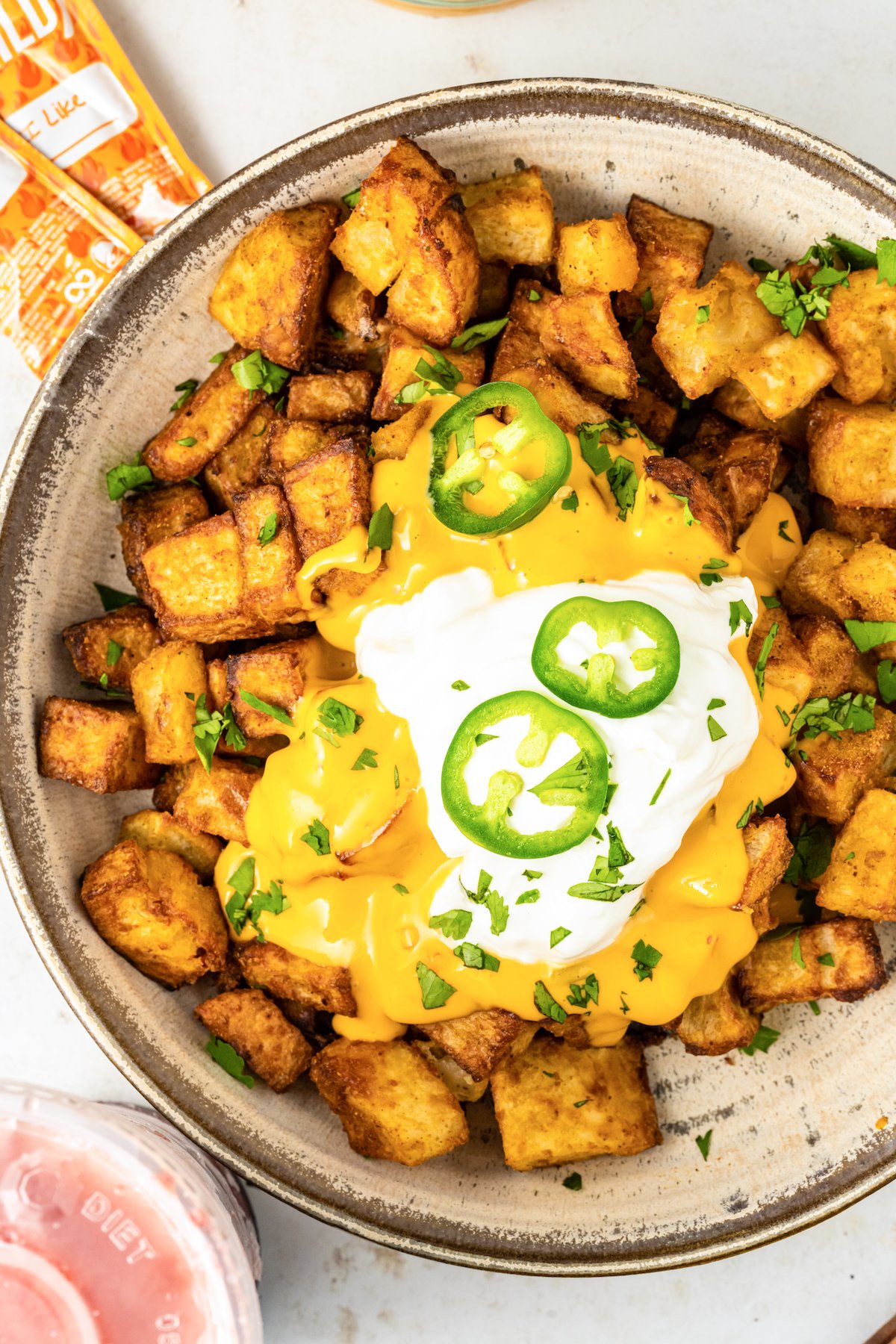A serving bowl filled with chopped seasoned fiesta potatoes covered in nacho cheese and sour cream. Slices of jalapeno and chives on top.