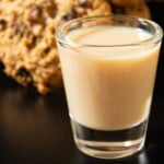 A shot glass filled with a creamy brown oatmeal cookie shot. Cookies are out of focus in the background.