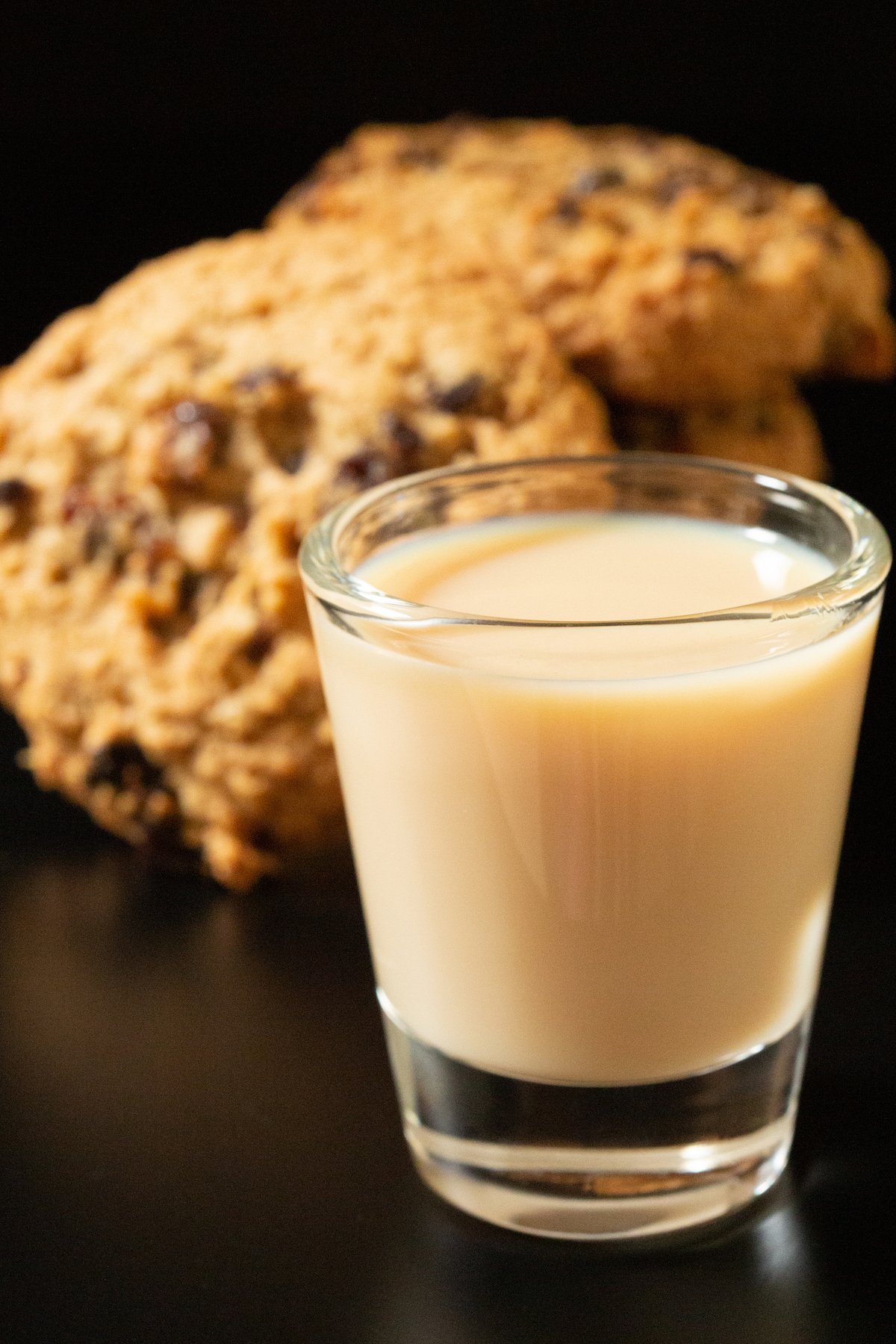 A shot glass filled with a creamy brown oatmeal cookie shot. Cookies are out of focus in the background.