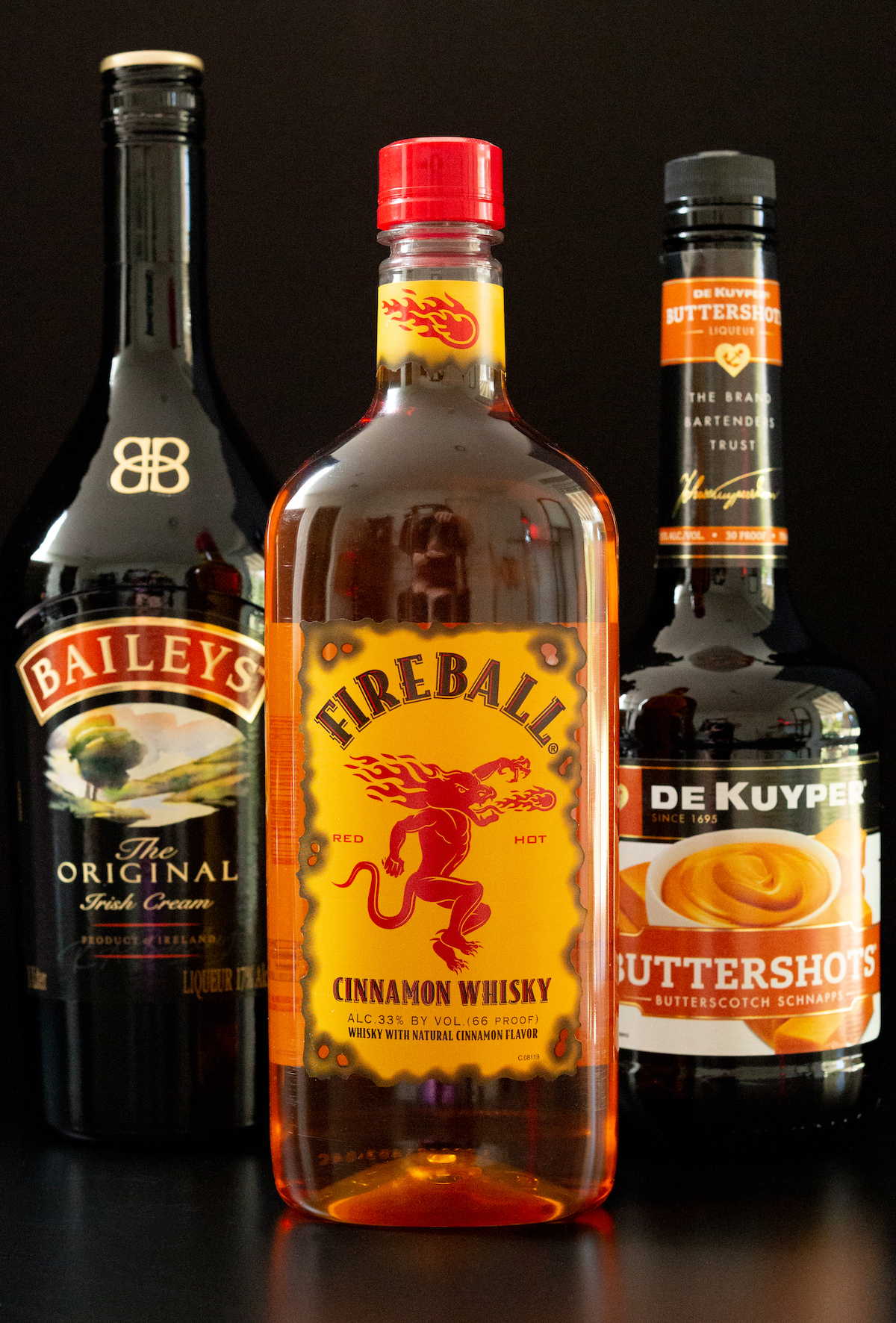 A bottle of Bailey's Irish Cream, Fireball, and Buttershots all on a black background.