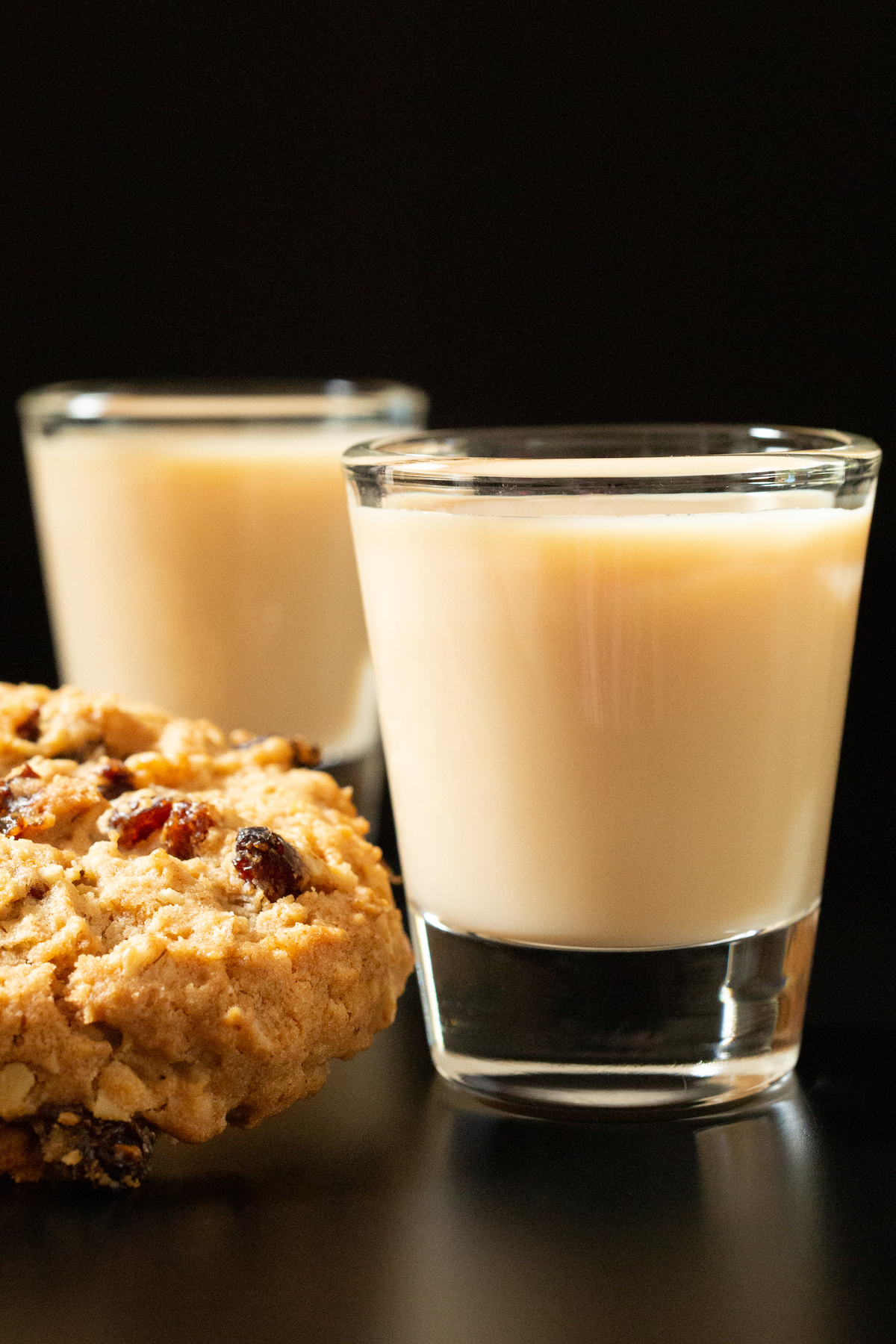 Two shot glasses filled with light brown oatmeal cookie shots next to a cookie on a black background.