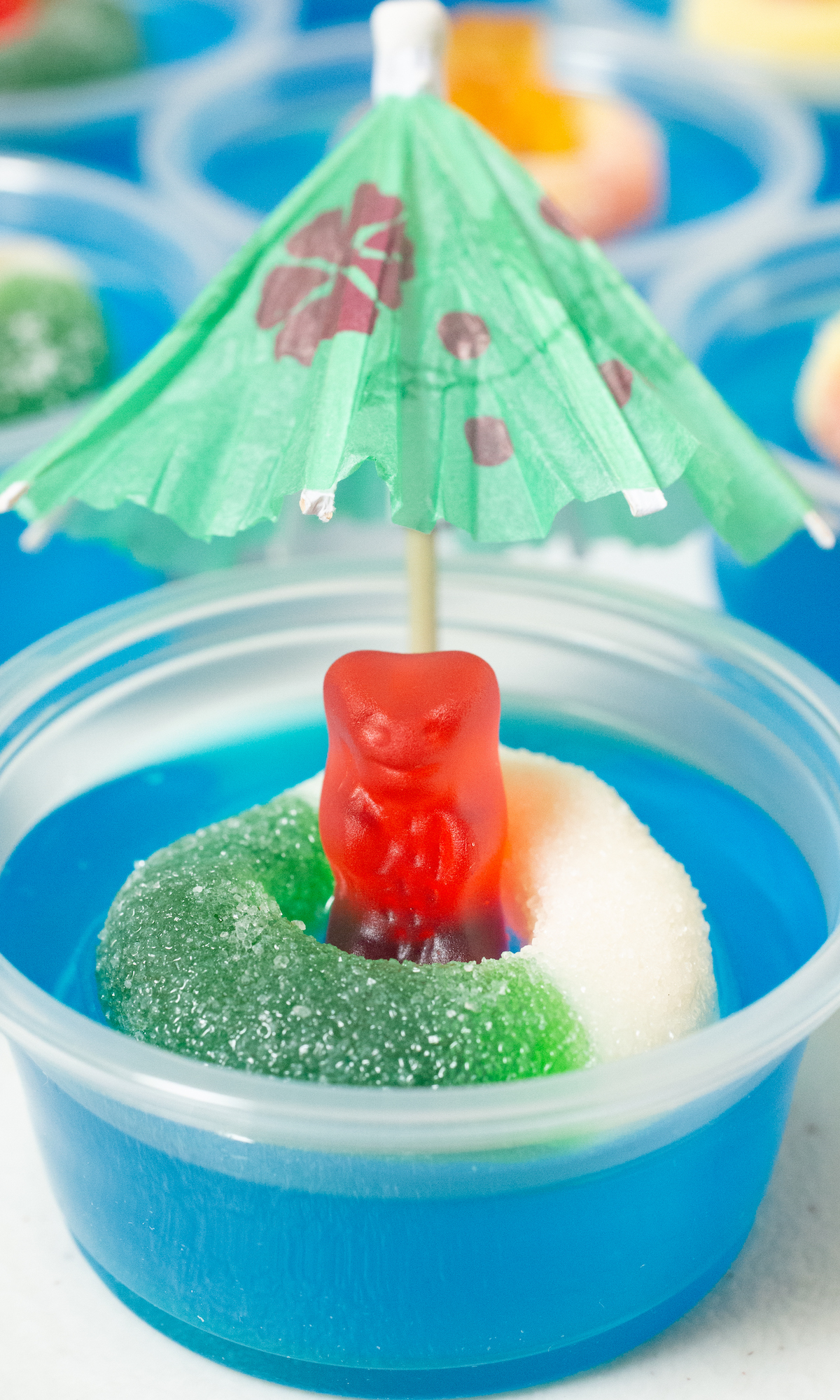 A single 2 oz plastic container filled with a blue jello shot that's topped with a gummy bear inside a gummy ring and garnished with a cocktail umbrella.
