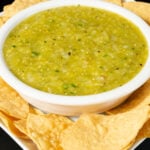 A white serving bowl filled with green Hatch chile on a serving plate surrounded by tortilla chips.