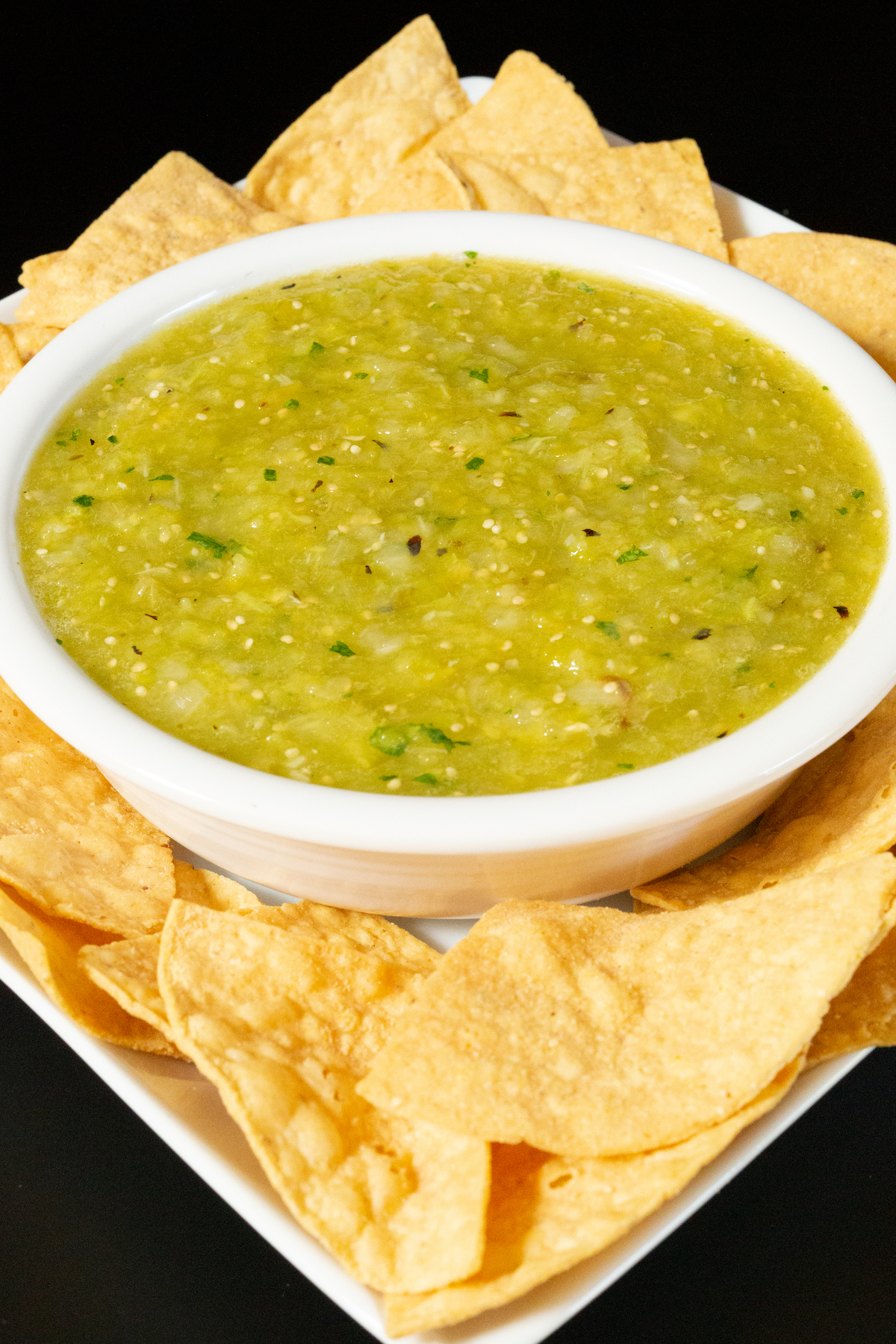 A white serving bowl filled with green Hatch chile on a serving plate surrounded by tortilla chips.