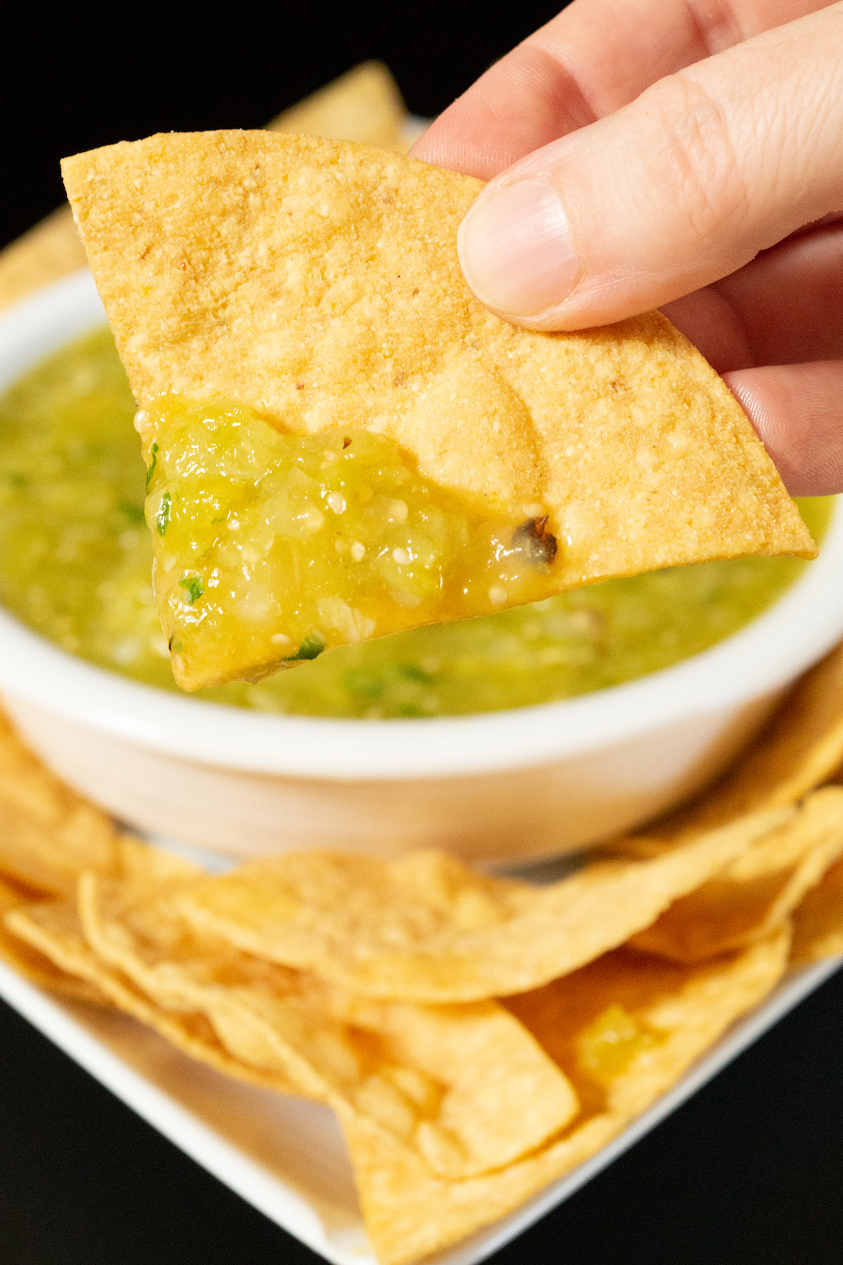 A hand holds up a single tortilla chip that's been dipped in green hatch chile salsa. A bowl with the rest is out of focus in the background.