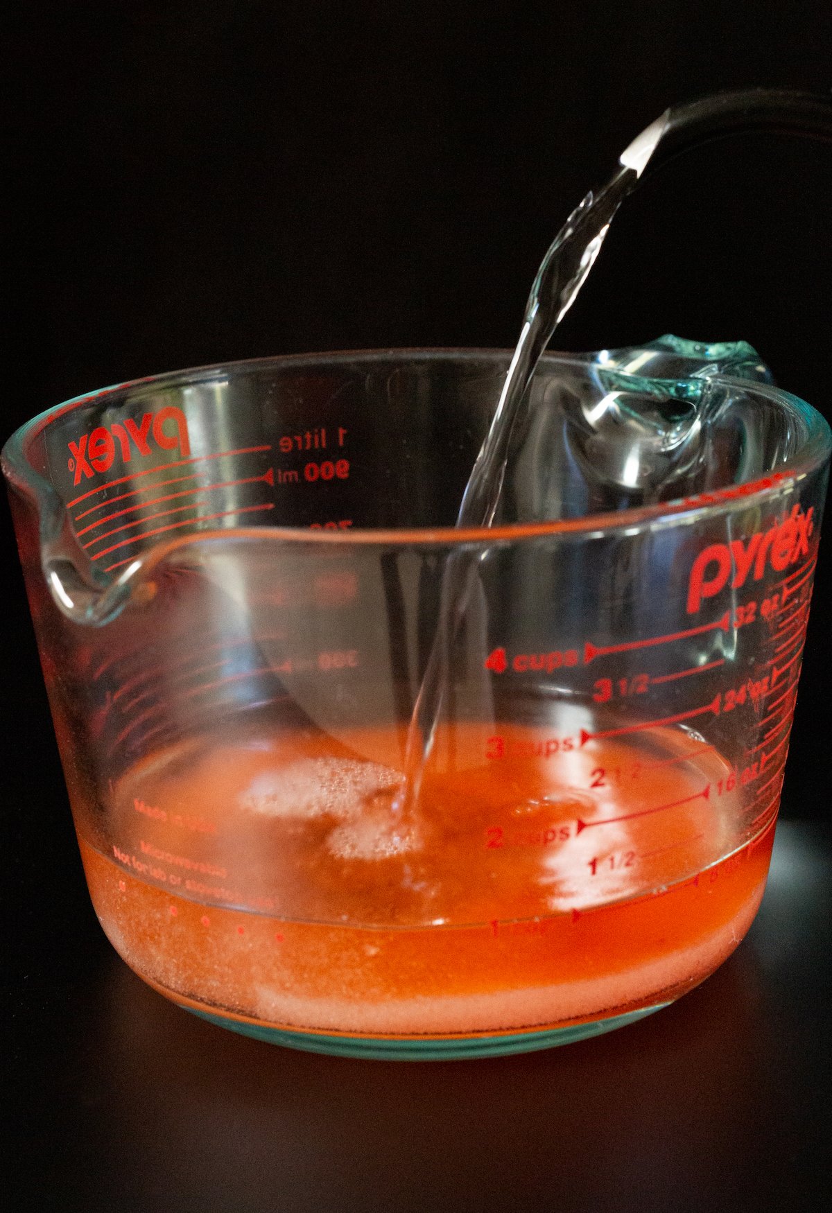 A large pyrex measuring cup with peach gelatin mix being filled with hot water from a kettle.