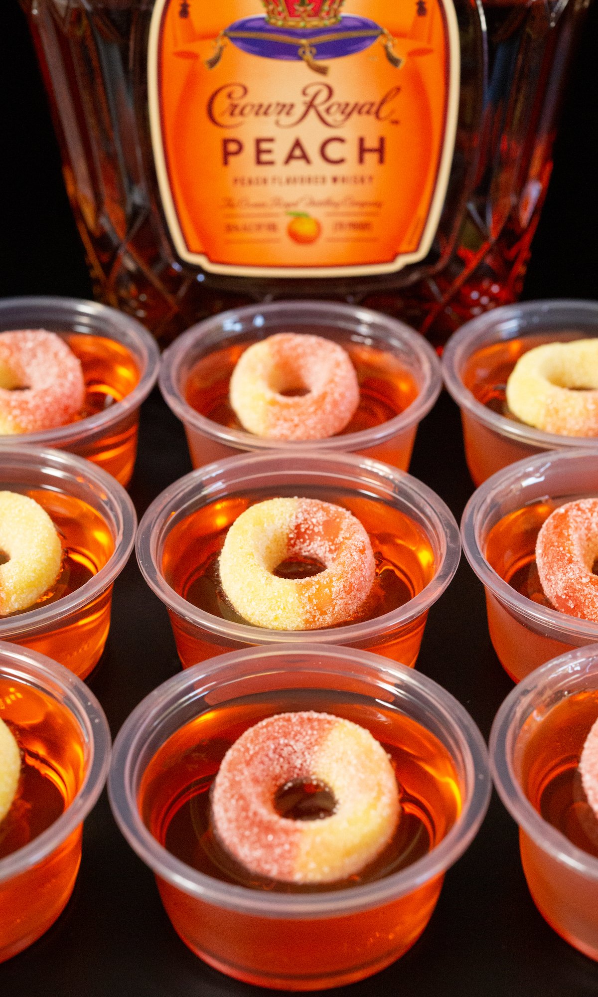 9 2 oz plastic containers filled with peach jello shots and topped with peach gummy candy rings in front of a Crown Peach bottle.