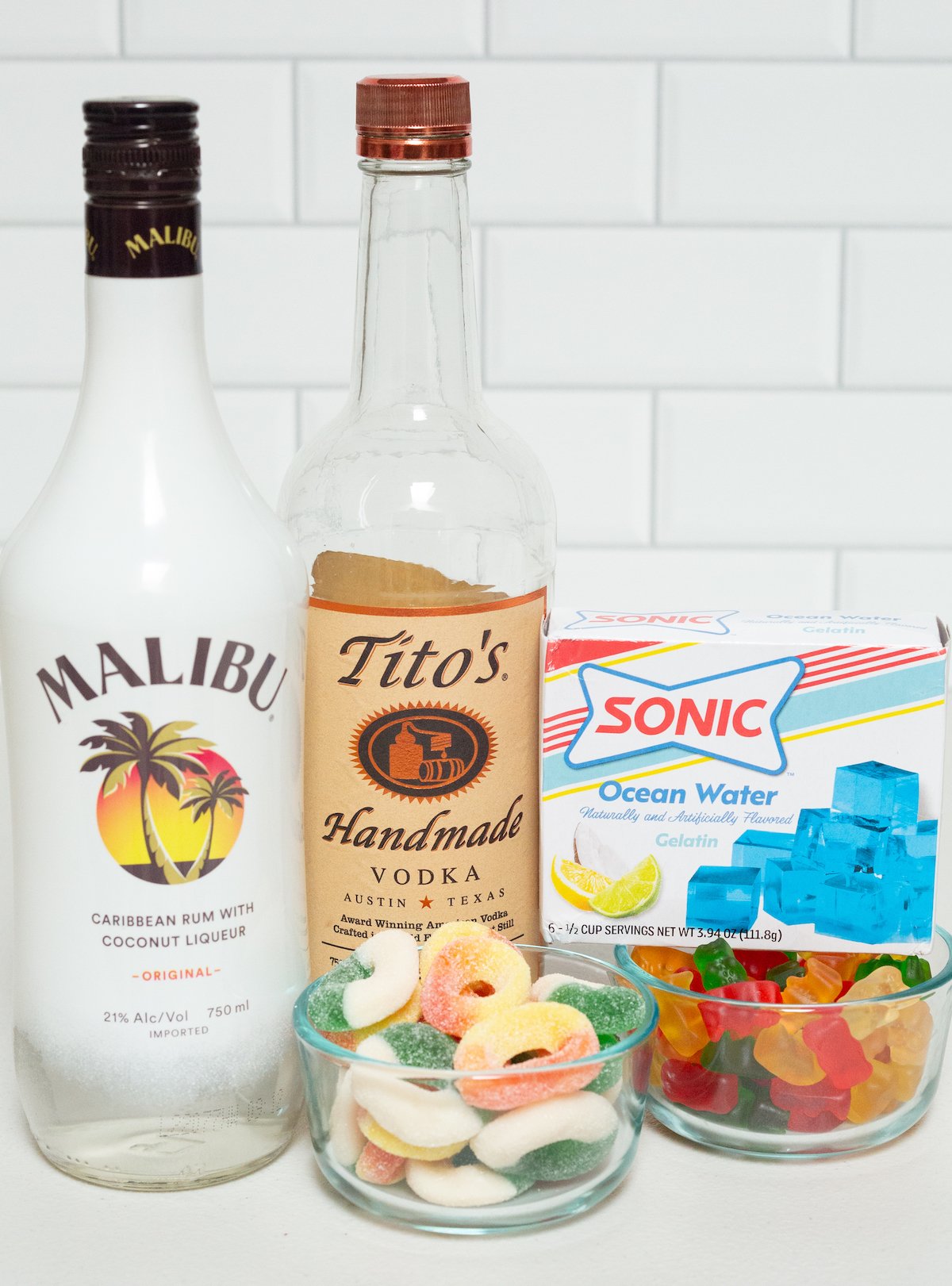 All the ingredients needed to make pool party jello shots on a white background - a bottle of Malibu & vodka, gelatin, gummy rings, and gummy bears.