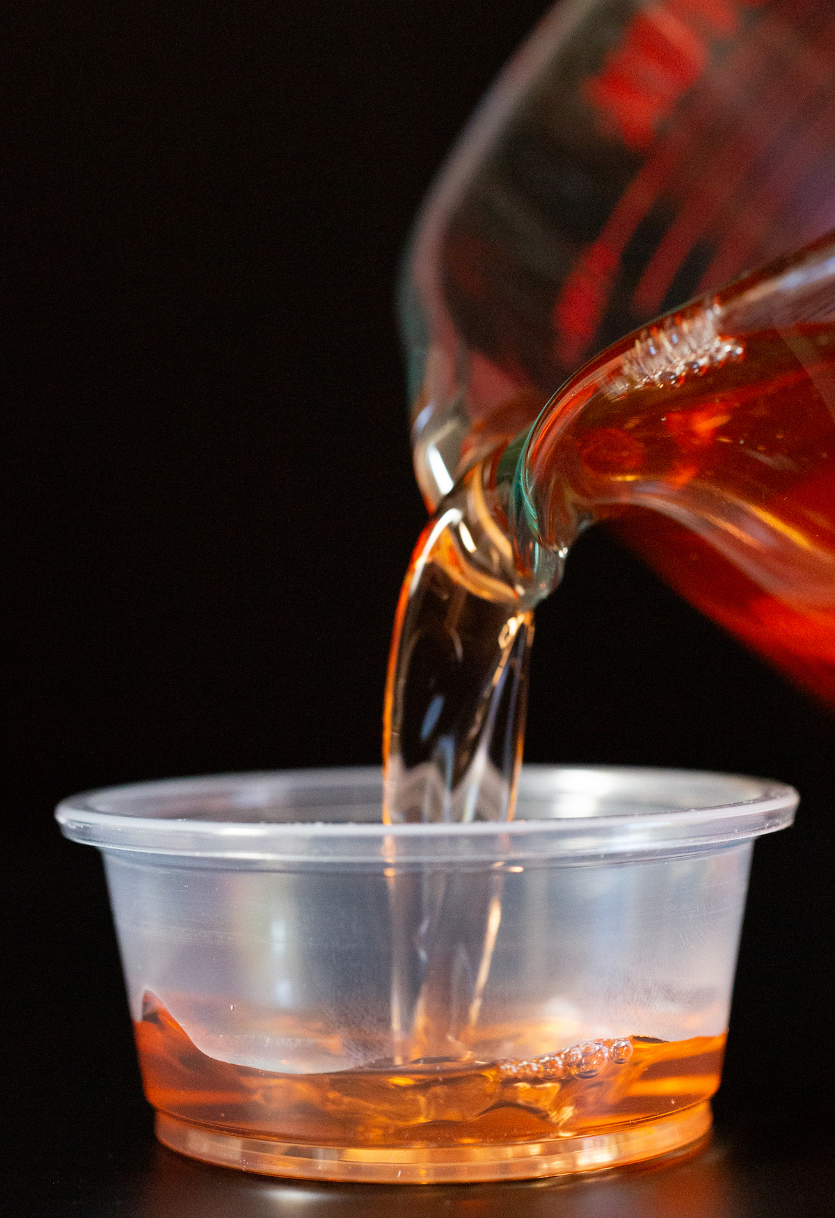 A measuring cup pours a peach jello shot liquid into a plastic 2 oz cup.