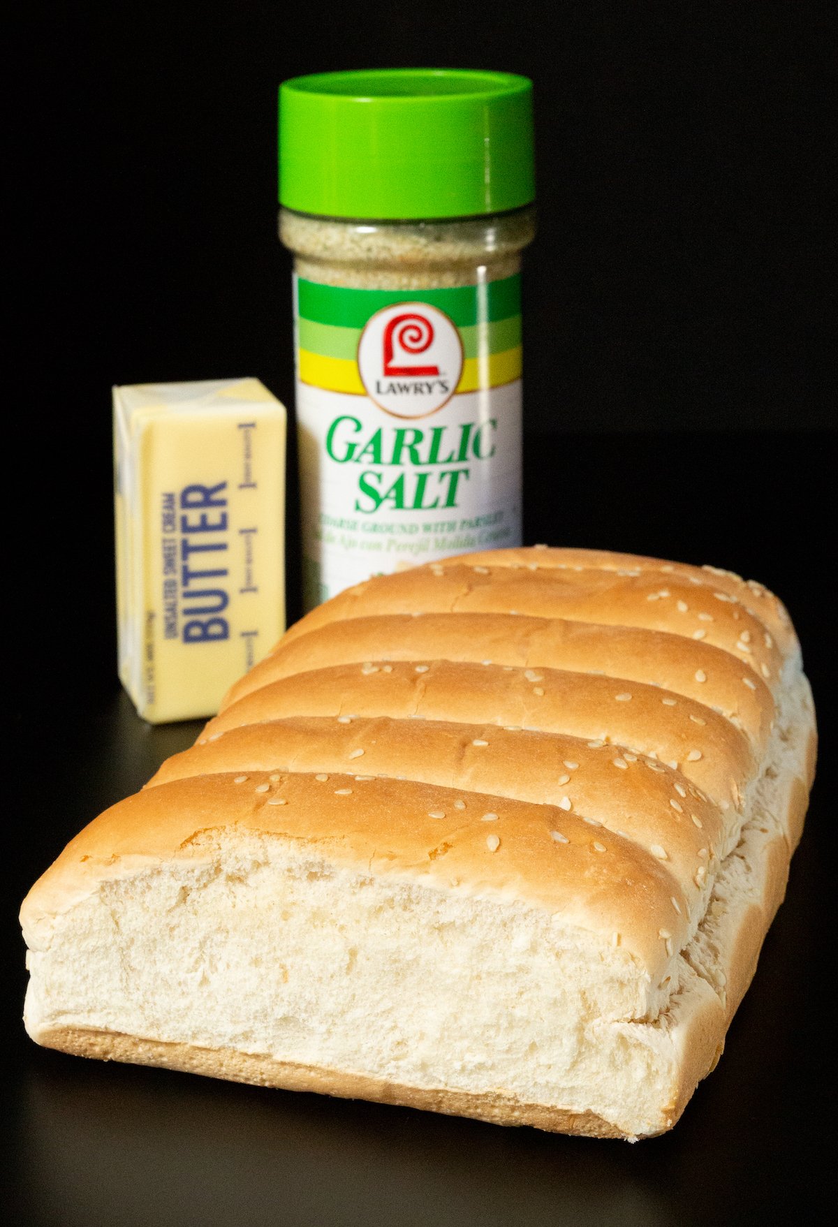 A stick of butter, a container of garlic salt, and a loaf of sesame bbq bread on a black background.