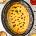 A crock pot that has a cooked dip that's topped with melted cheese, diced tomato, sliced black olives, and chopped cilantro.