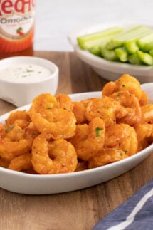 A white serving dish filled with buffalo shrimp. Hot sauce, ranch, and celery sticks out of focus in background.