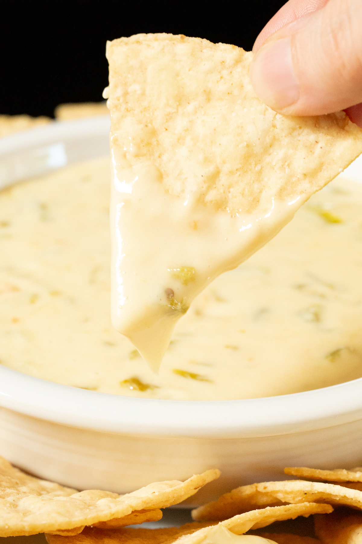 A hand holds up a tortilla chip that has been dipped in hatch chile queso. A bowl with the rest of the dip is out of focus in the background.