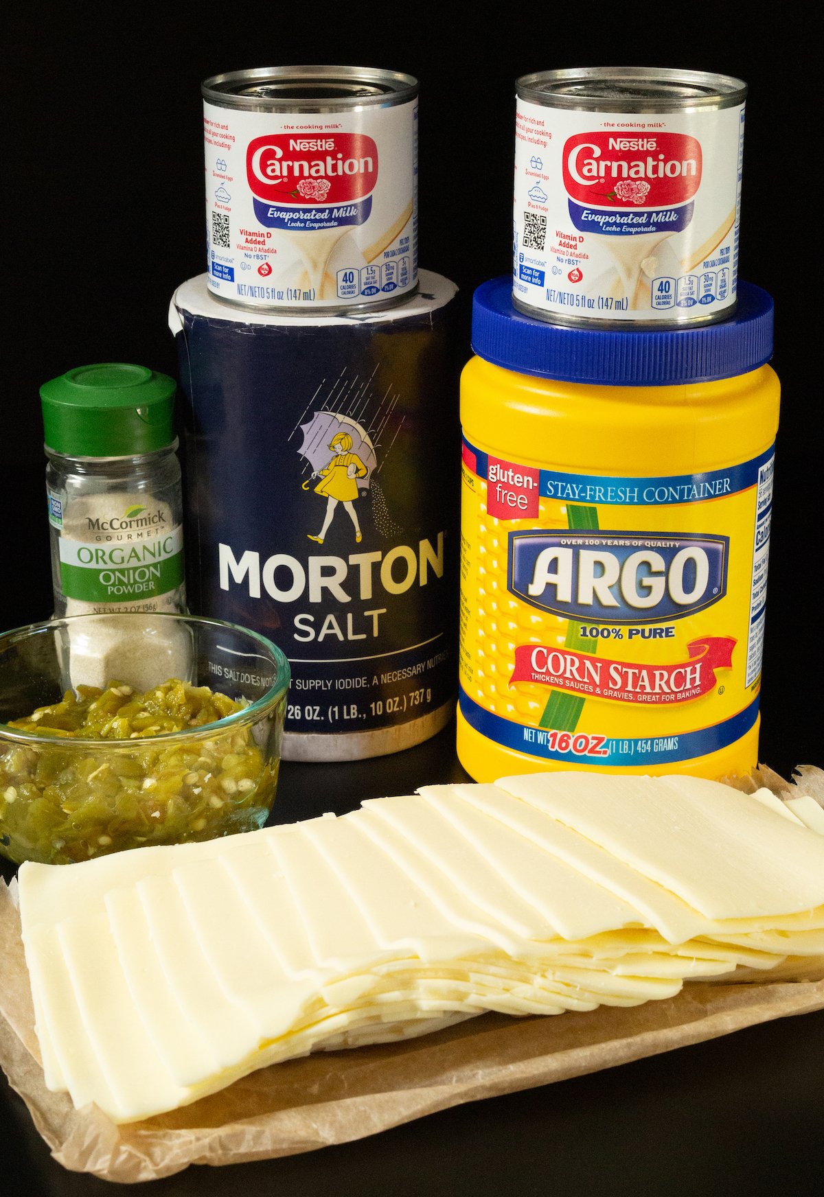 A small prep dish of chopped hatch chiles and slices of white American cheese next to containers of cornstarch, salt, onion powder, and evaporated milk.