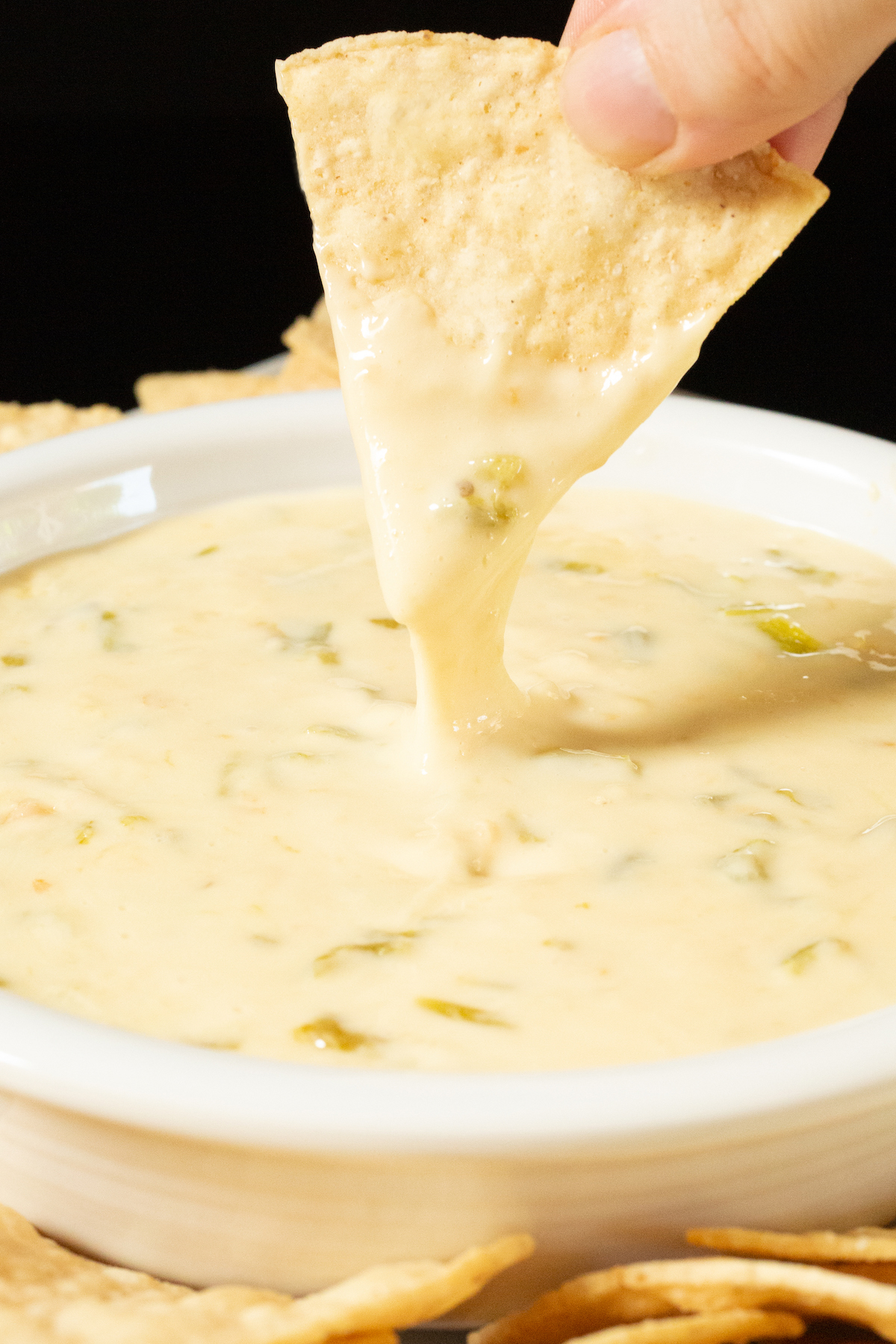 Two fingers pull a tortilla chip up from being dipped in a bowl of hatch chile queso, the cheese dip stretches with the chip.