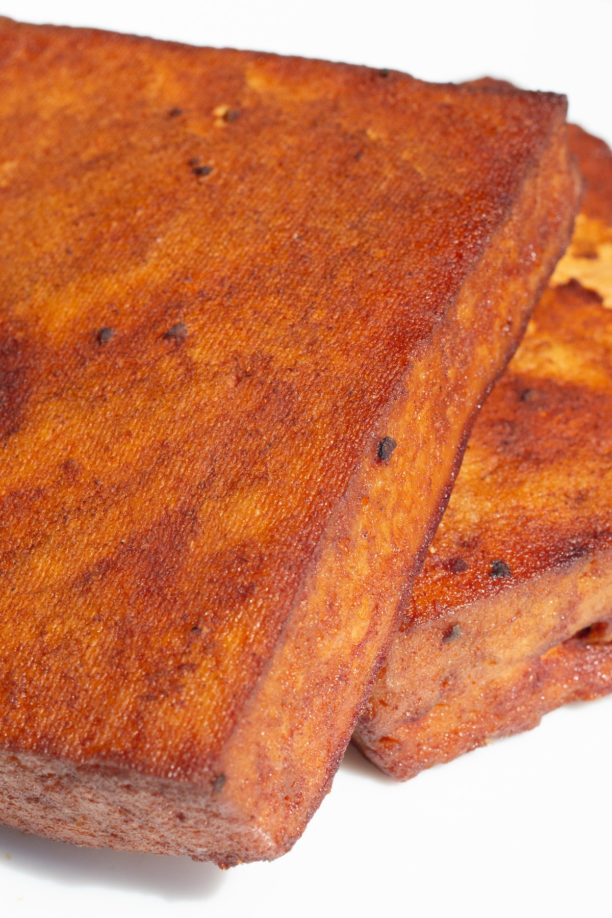 Two blocks of tofu that have turned deep red from marinating in bbq sauce and being smoked on the grill on a white serving plate.