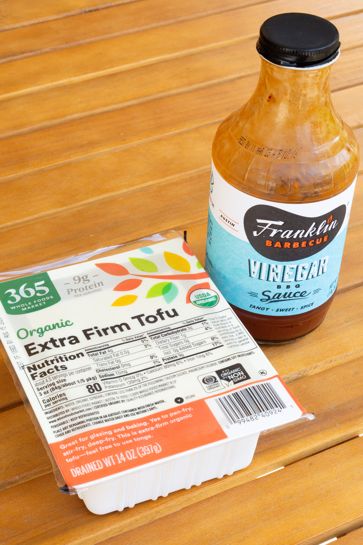 A package of extra firm tofu and a bottle of bbq sauce on a wooden outdoor table.