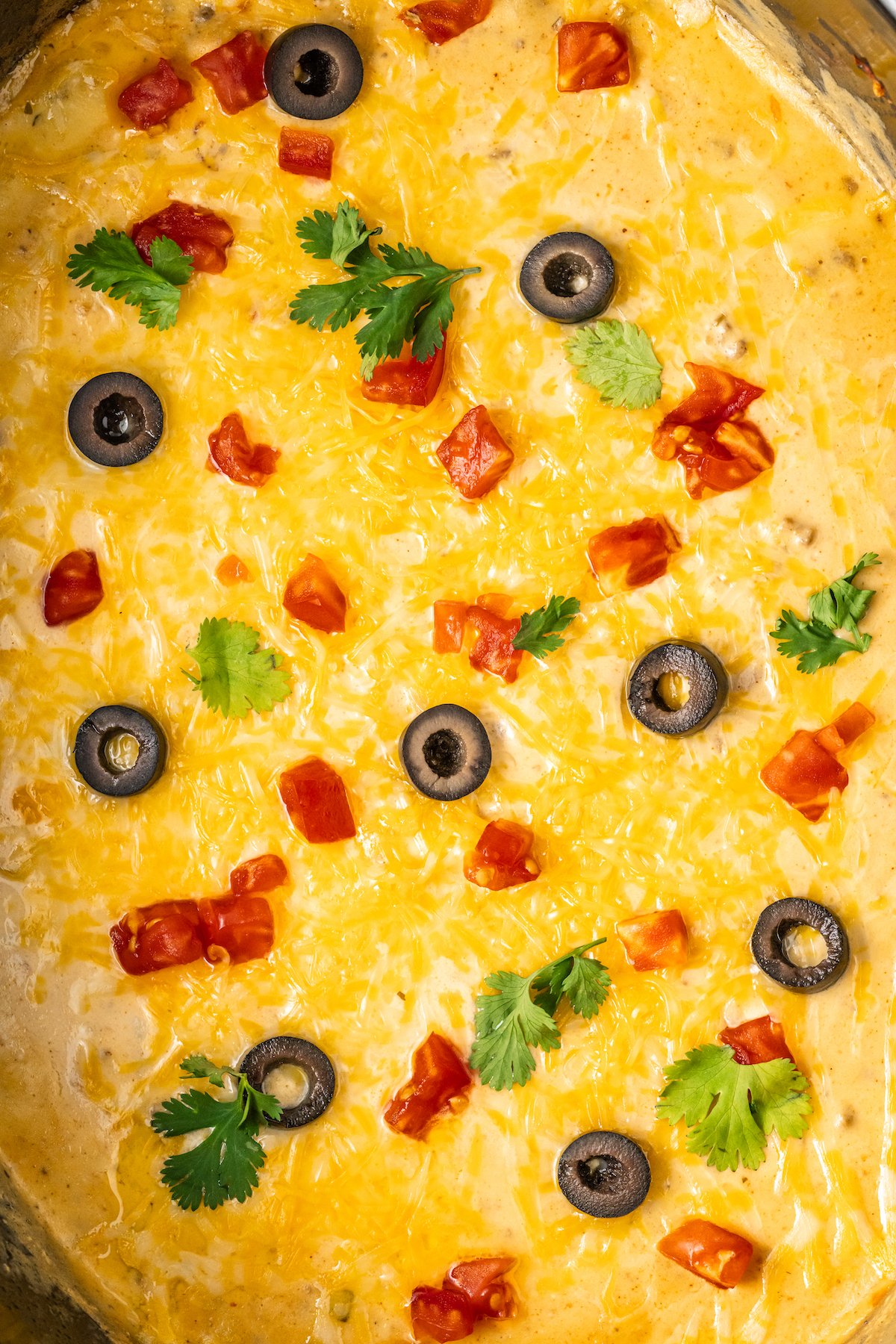 Up close view of the top of the taco dip in the crock pot featuring the shredded melted cheese, sliced olives, chopped tomato and cilantro.