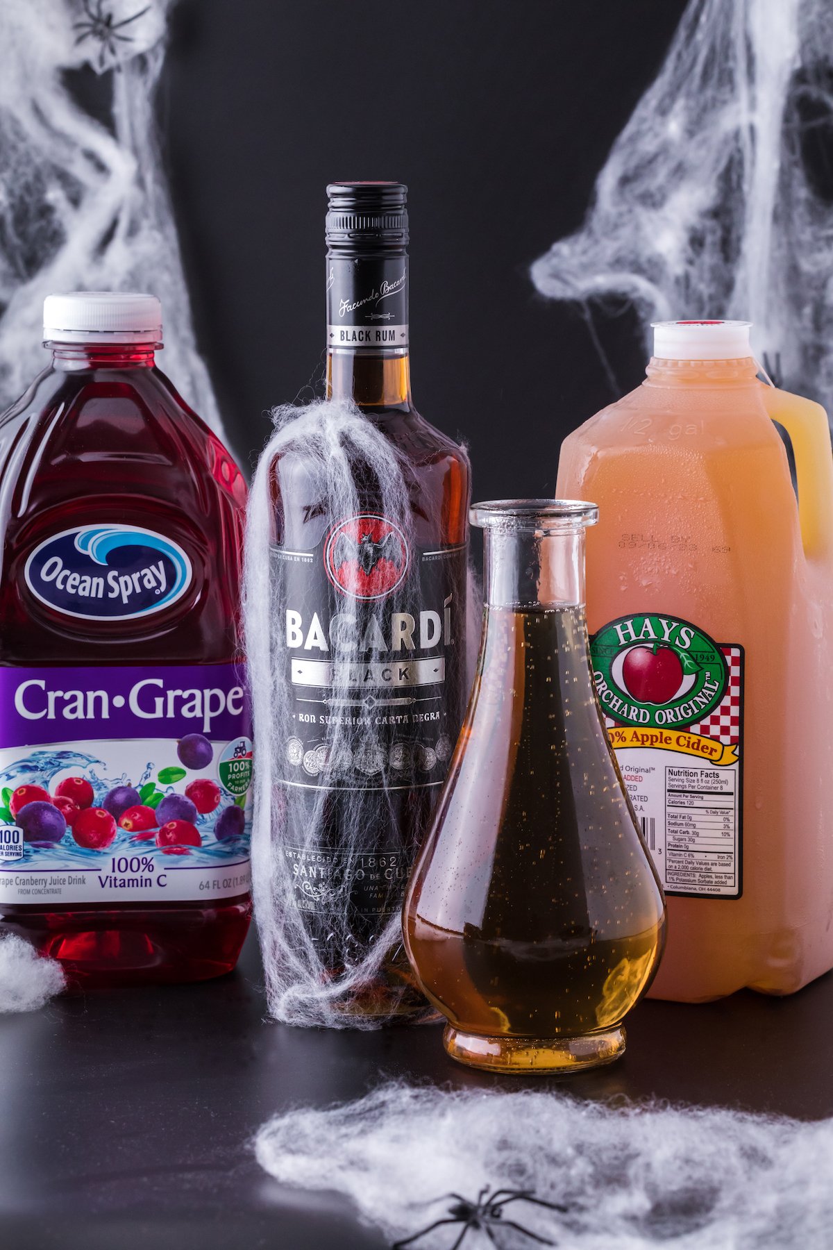 A bottle of Cran-Grape juice, Bacardi dark rum, apple cider, and ginger on a black background.