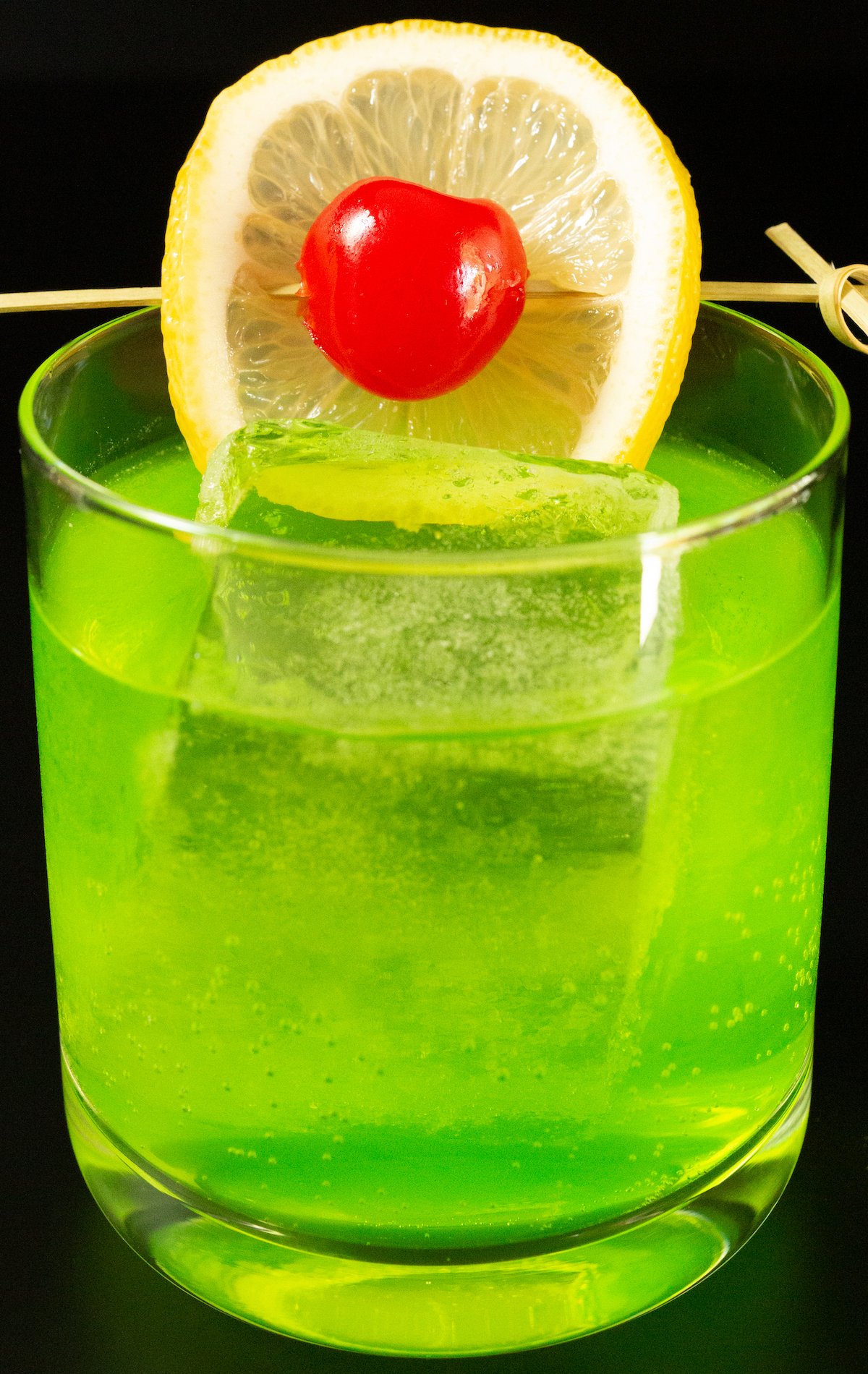 A lowball glass with a large block of ice is filled with a bright neon green Midori Sour cocktail that's garnished with a lime wheel and a maraschino cherry on a skewer.