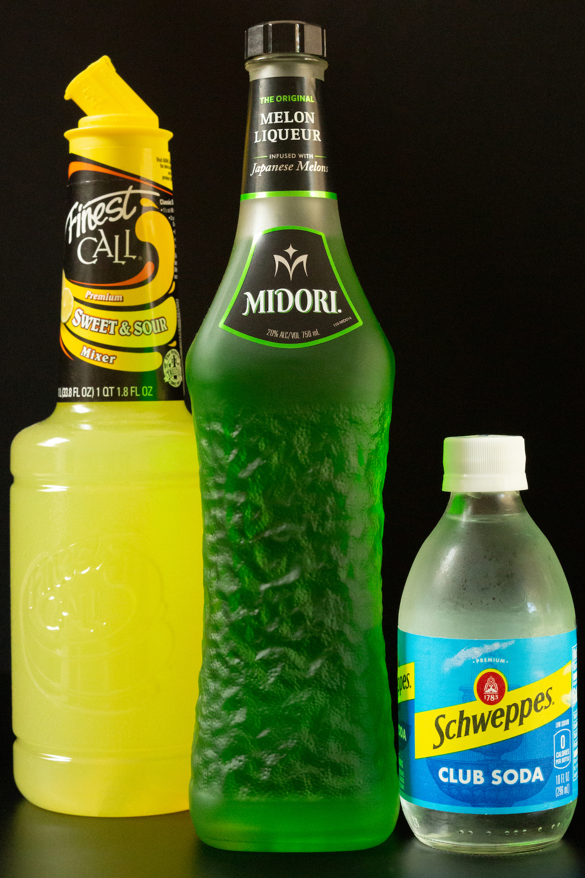 A bottle of sweet 7 sour mix, Midori, and club soda all on a black background.