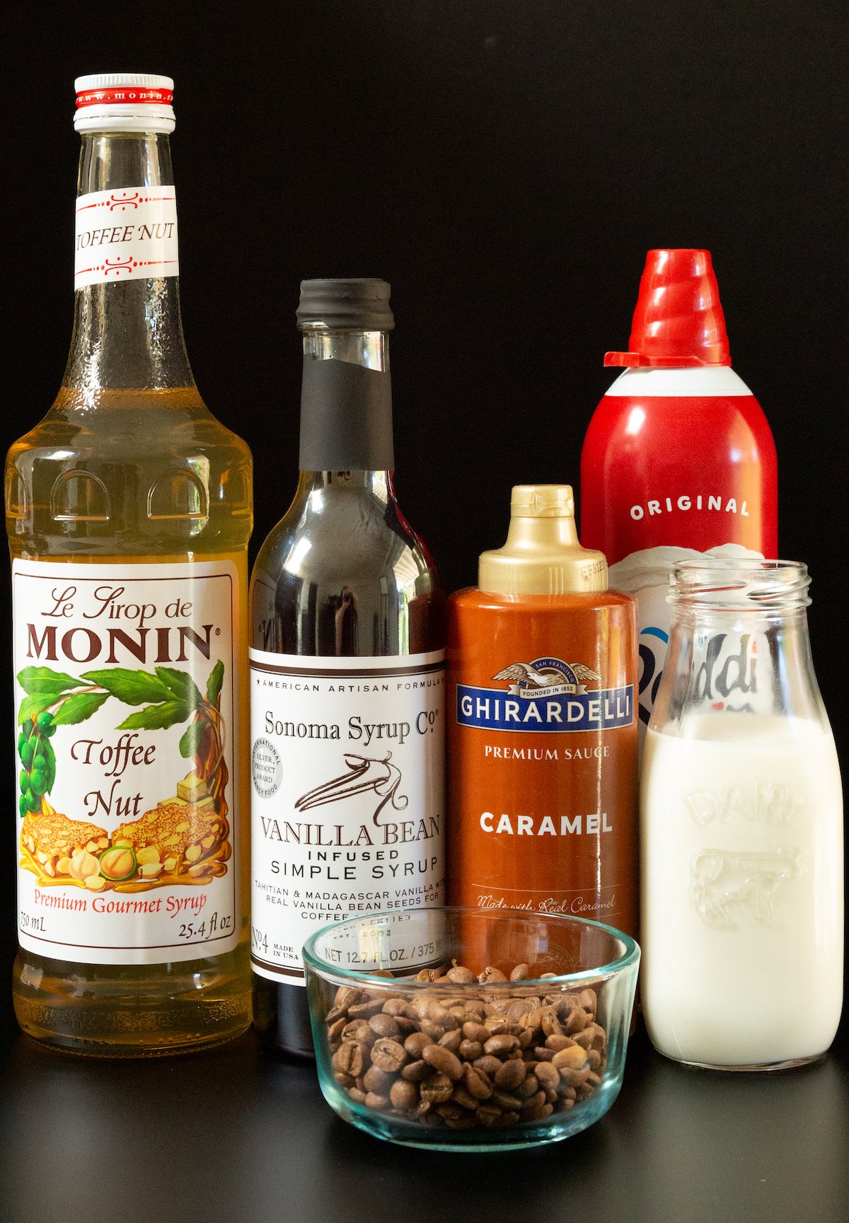 A bottles of toffee nut syrup, vanilla bean syrup, caramel, milk, and whipped cream on a black background next to a small bowl of coffee beans.