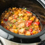 A crock pot is filled with a mixture of cooked chicken thighs, bell peppers, pineapple chunks, and green onions in a thick, sweet brown sauce.