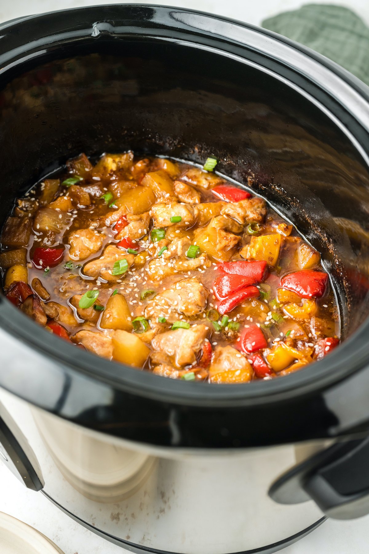 A crock pot is filled with a mixture of cooked chicken thighs, bell peppers, pineapple chunks, and green onions in a thick, sweet brown sauce.