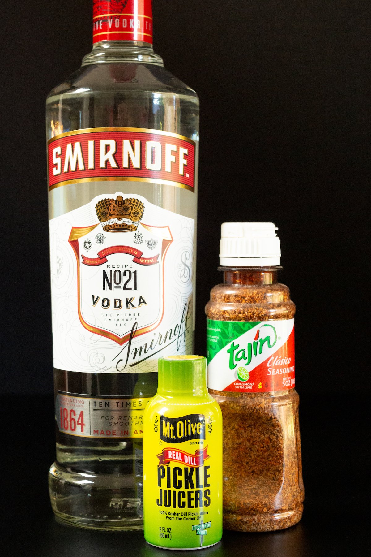 All the ingredients to make pickle shots on a black background - a bottle of vodka, Tajin, and pickle juice.