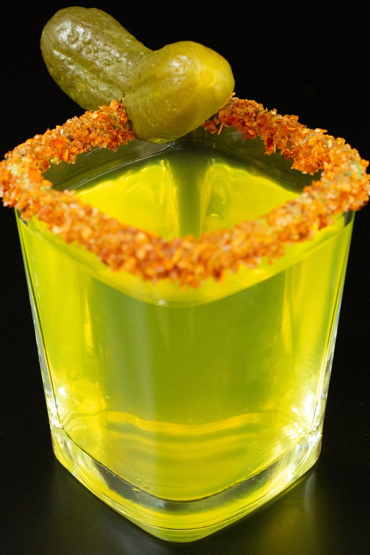 A single square shot glass filled with a neon green pickle shot. The rim has been dipped in red Tajin chili powder and a baby pickle is added as a garnish.