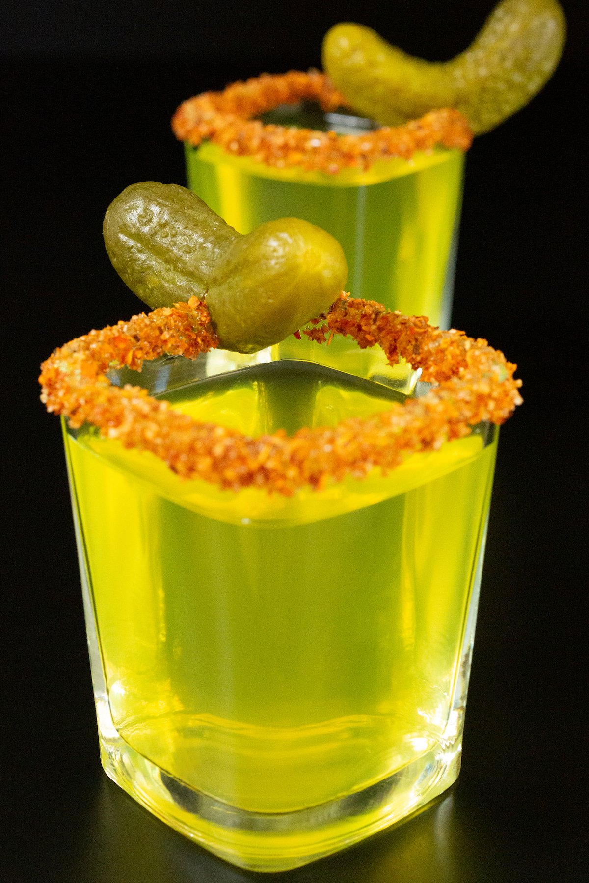 Two square shot glasses filled with neon green pickle shots garnished with a Tajin rim and a baby pickle.