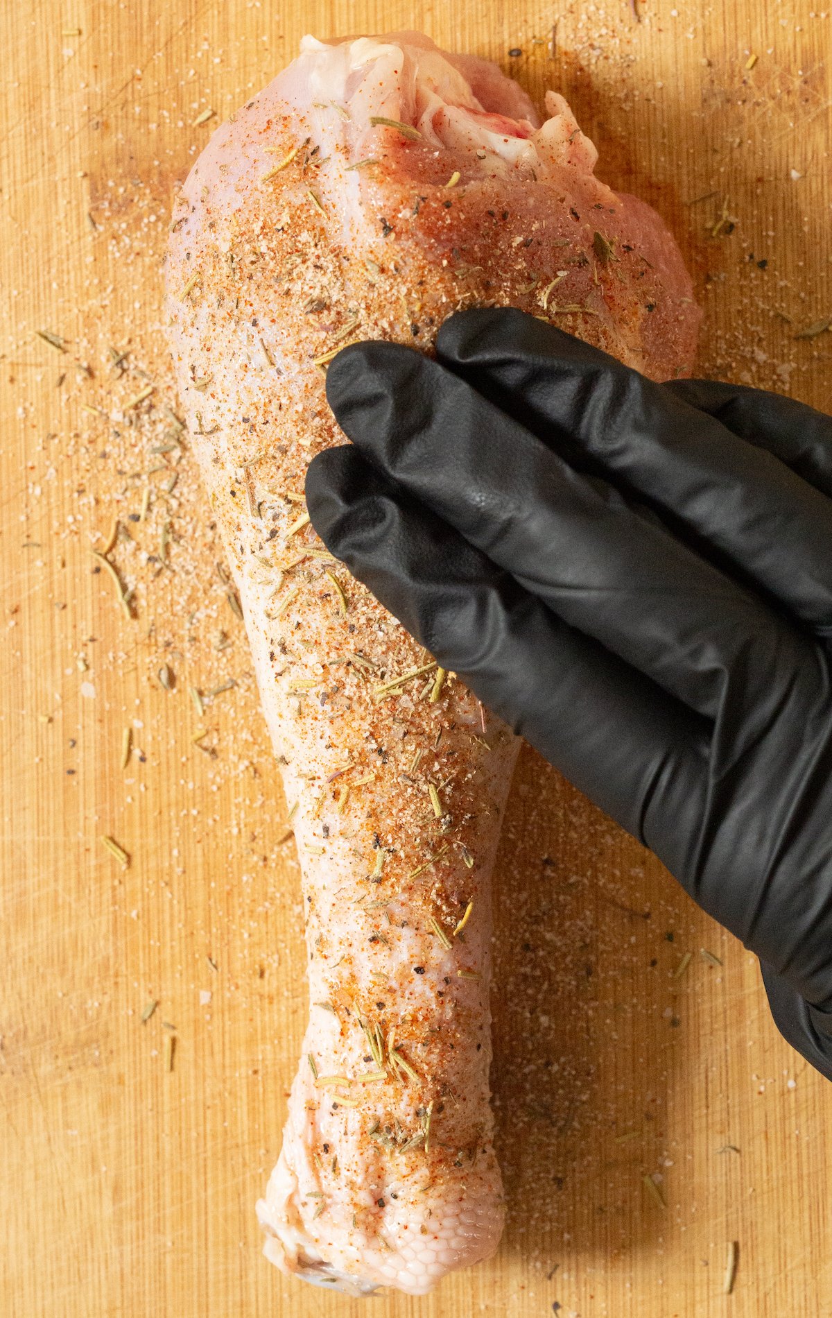 A raw turkey leg covered in seasoning that's being rubbed in by a gloved hand.
