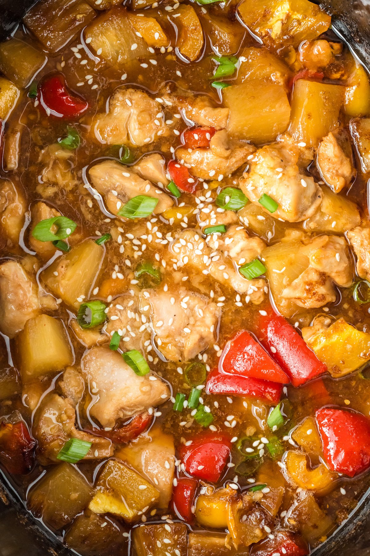 Close up of hawaiian chicken made in a crock pot featuring diced chicken thighs, diced bell peppers, pineapple chunks in a thick brown sauce. Garnished with sesame seeds and green onion.