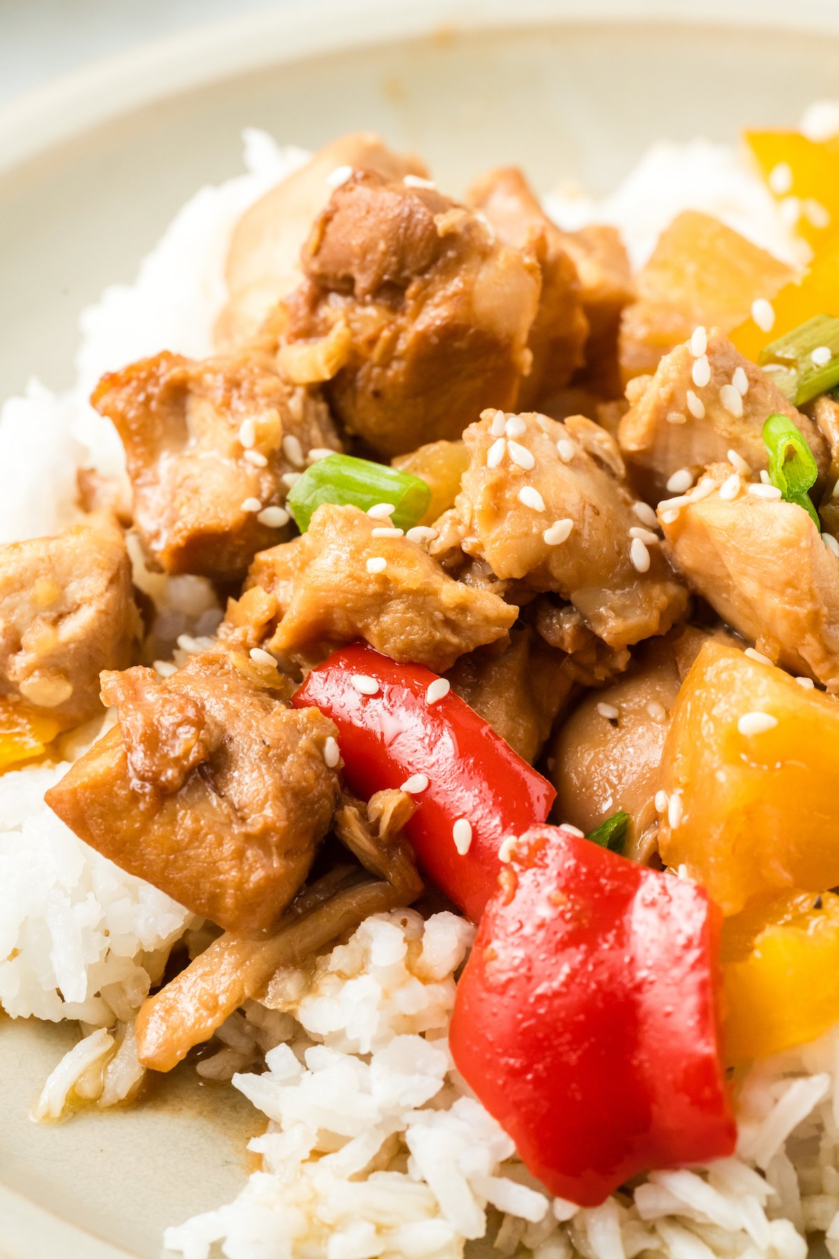 Close up of sweet hawaiian chicken thighs with red bell pepper and pineapple chunks on top of white rice on a dinner plate.