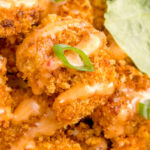 Close up of a single breaded fried shrimp that's drizzled in a orange colored bang bang sauce and garnished with a sliced green onion.