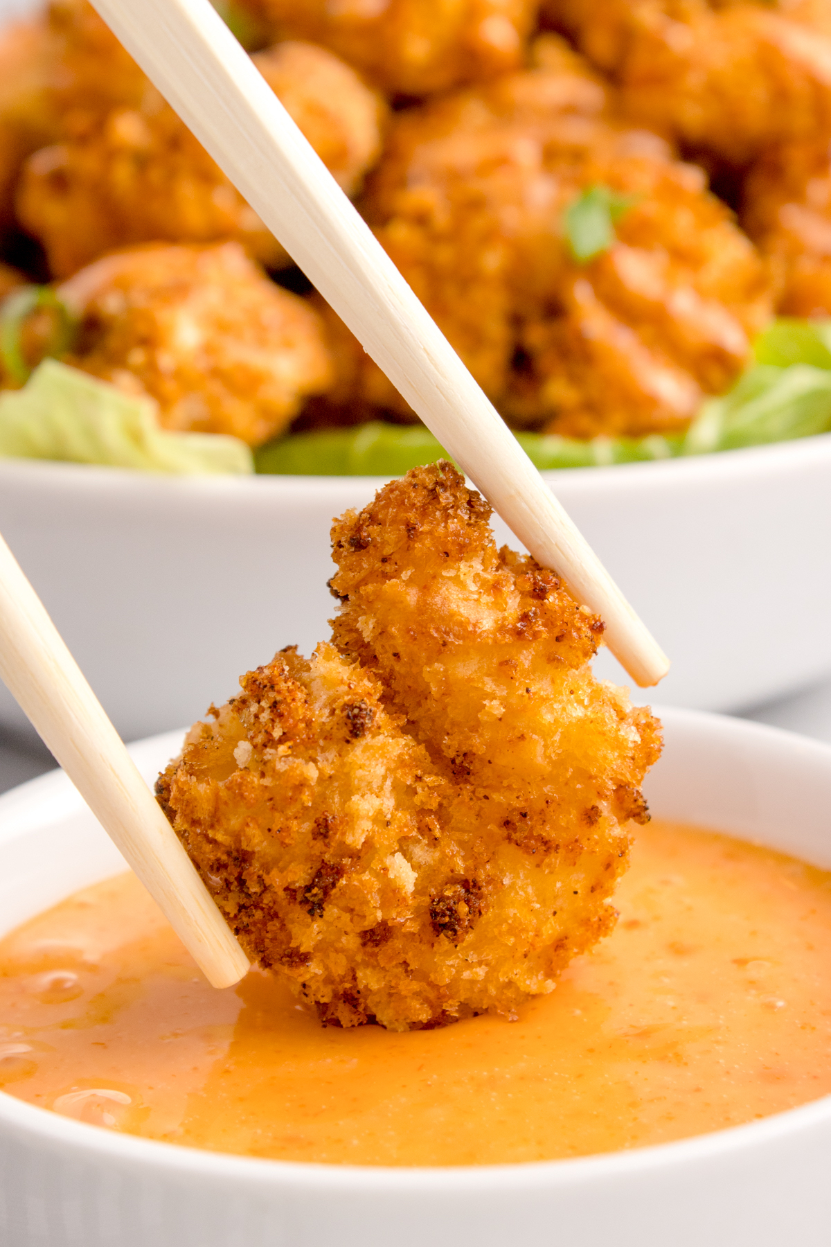 A pair of chopsticks holds a crispy breaded shrimp that's being dipped in a bowl or orange colored bang bang sauce.