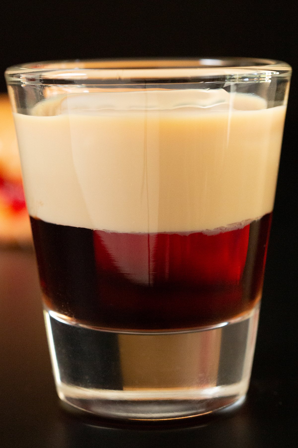 A shot glass is filled with two distinct layers - a crimson red on the bottom, and a creamy beige liqueur on top.