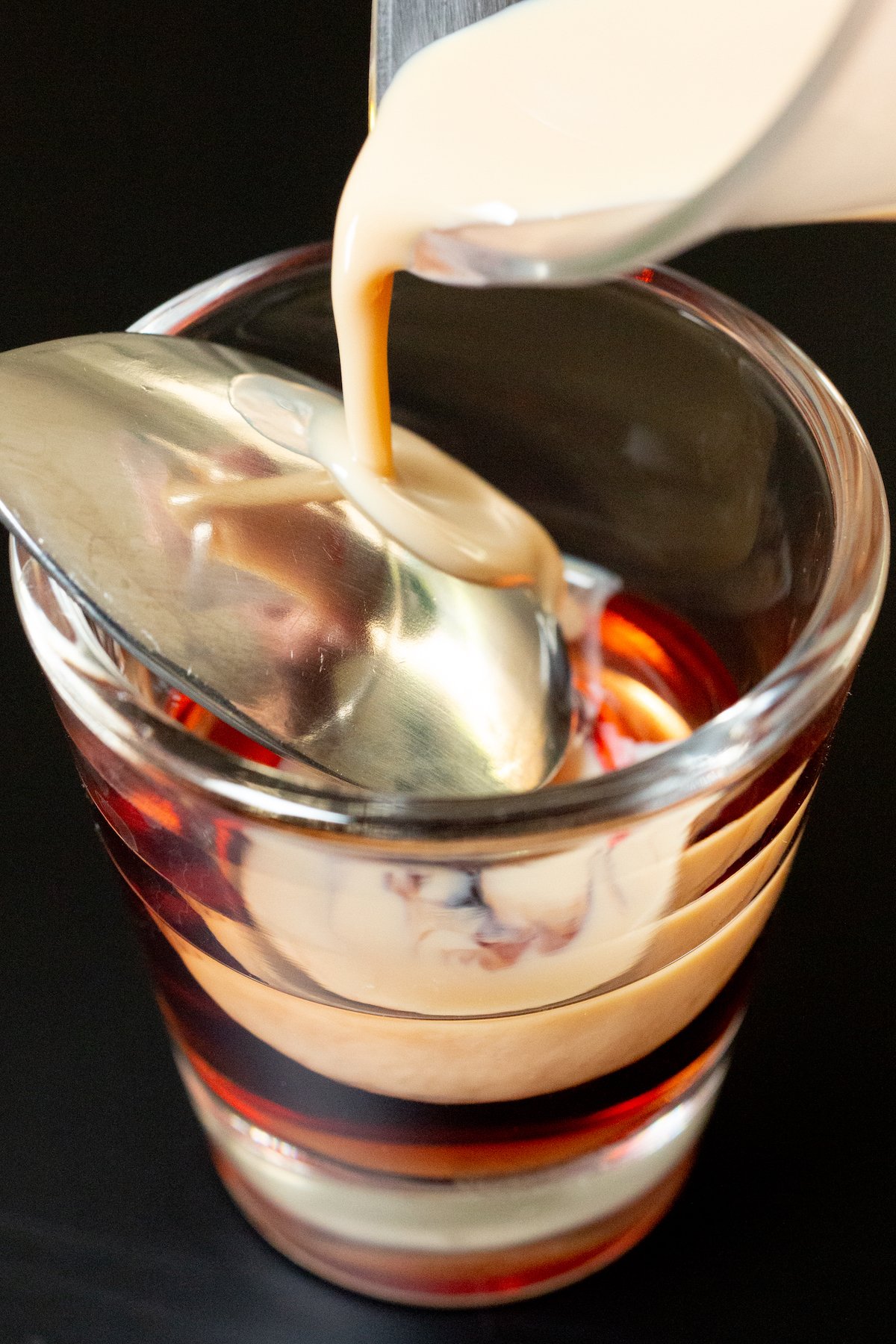 Irish cream being slowly poured over the back of a metal spoon that's positioned just above a layer of Chambord in a shot glass. 