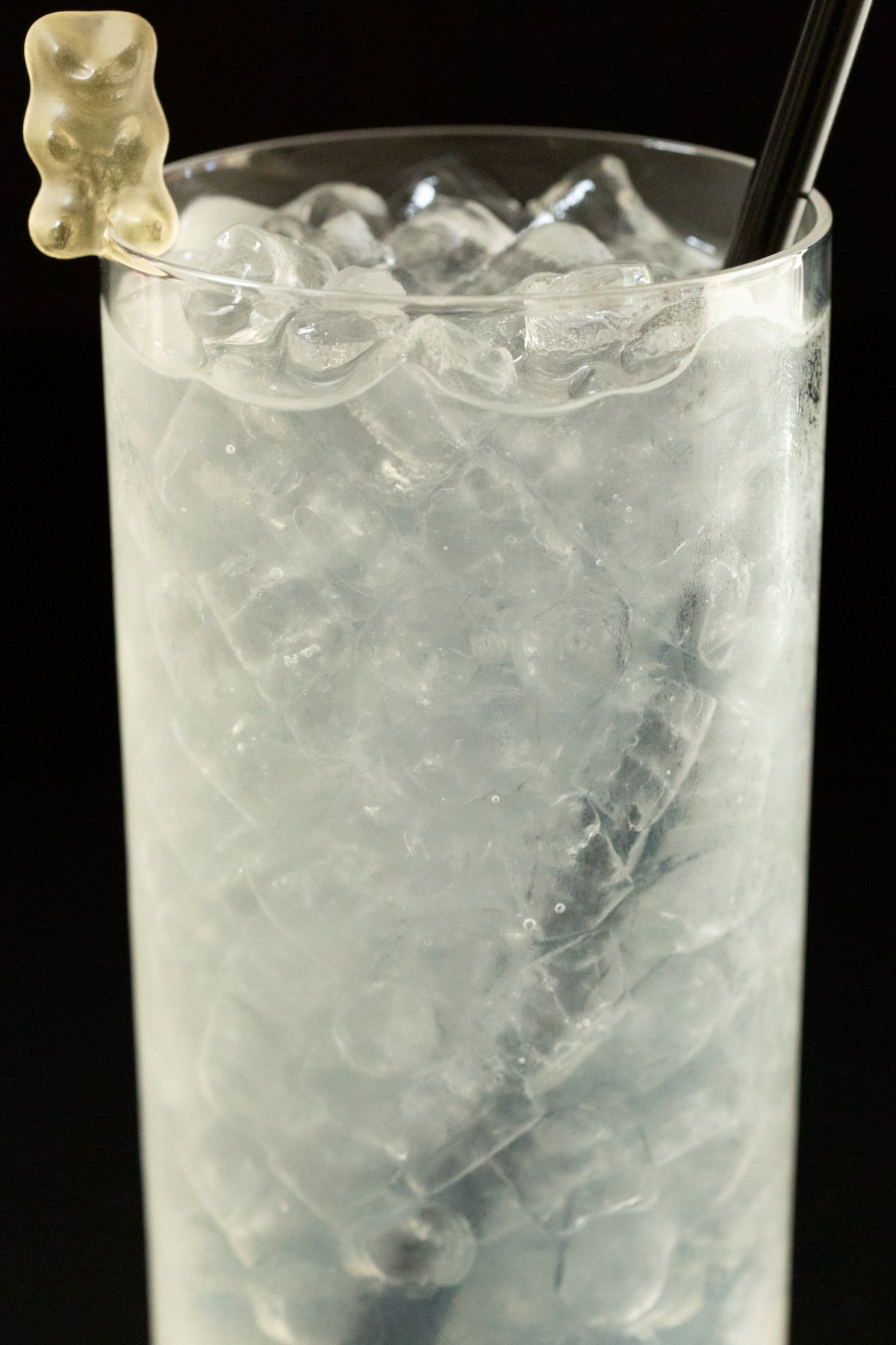 A tall glass is filled with pebble ice and white colored cocktail that's garnished with a white gummy bear.