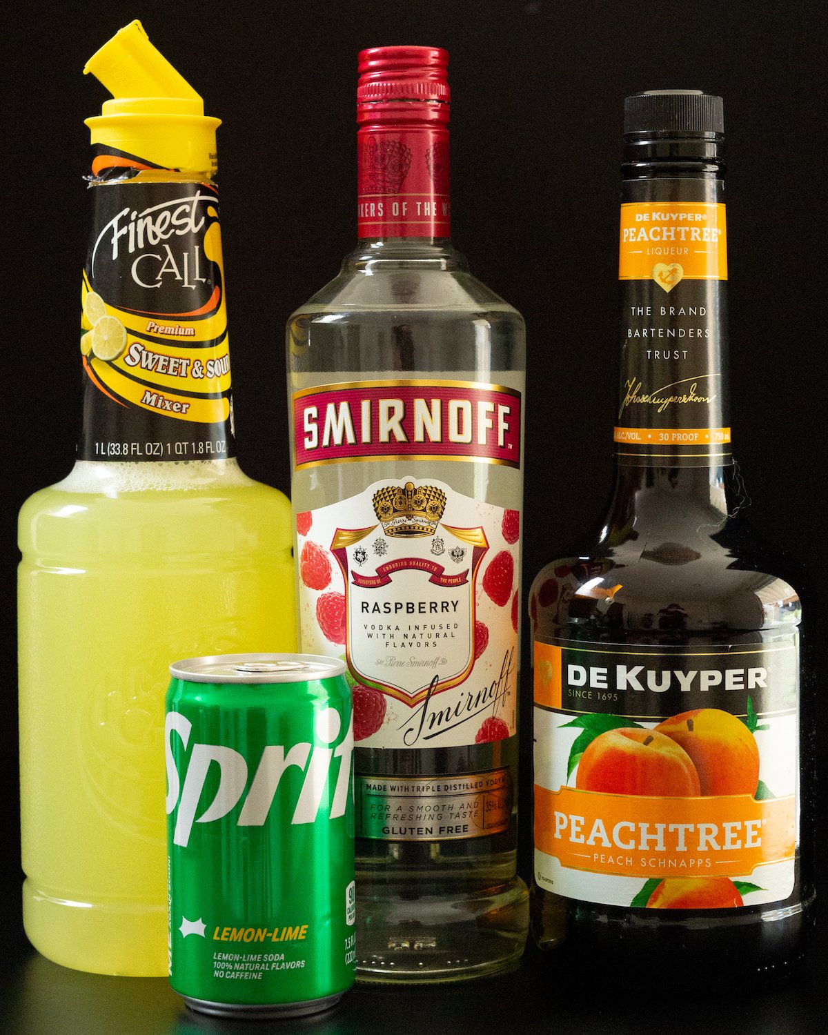 A bottle of sweet and sour mix, raspberry vodka, peach schnapps, and a can of Sprite on a black background.