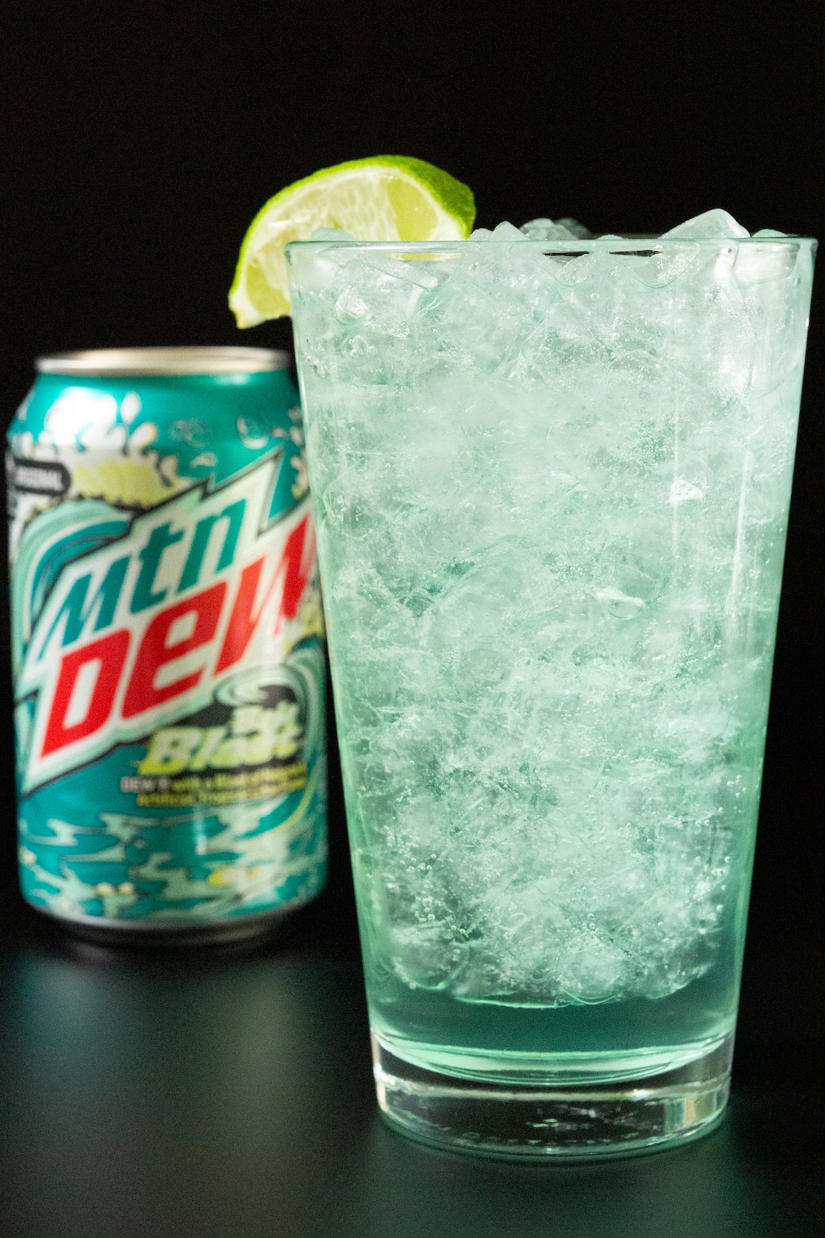 A pint glass filled with pebble ice and a turquoise colored cocktail. A can of Baja Blast is out of focus in the background.