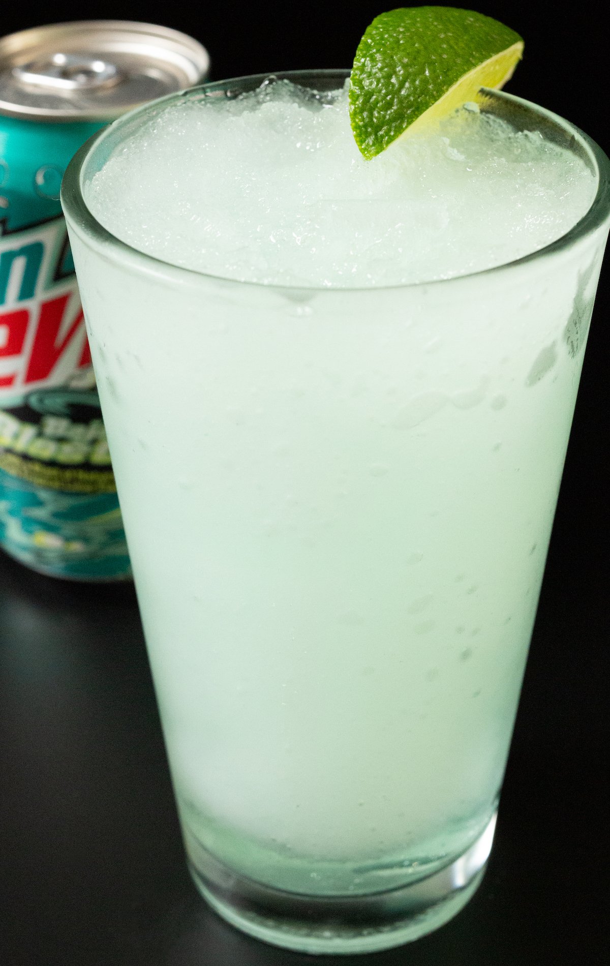 A pint glass is filled a a frozen blended light blue colored Baja Blast cocktail that's garnished with a lime wedge.