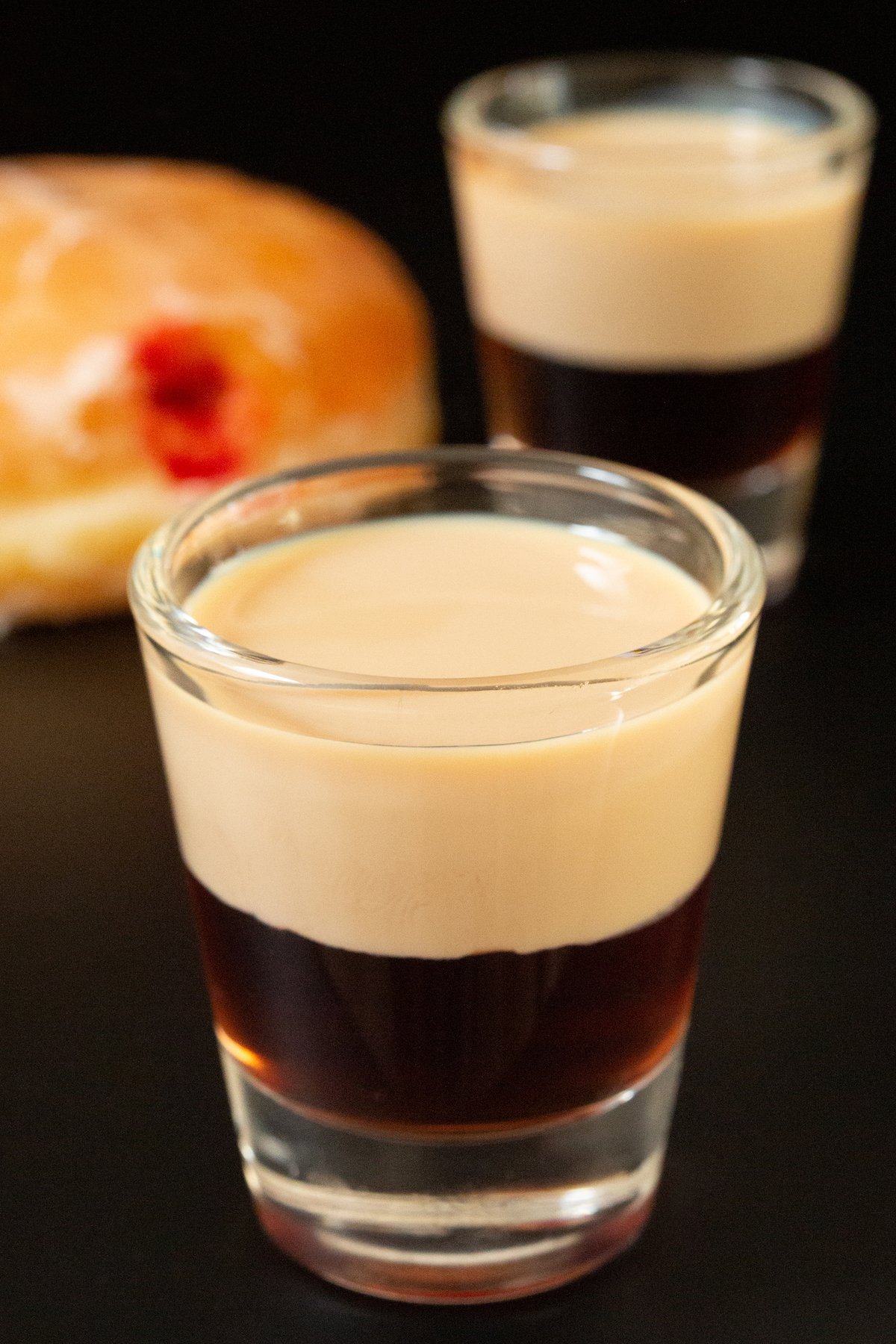 A shot glass filled with a layer of raspberry liqueur and a top layer of Irish cream is in focus in the foreground. Another is out of focus in the background next to a jelly donut. 