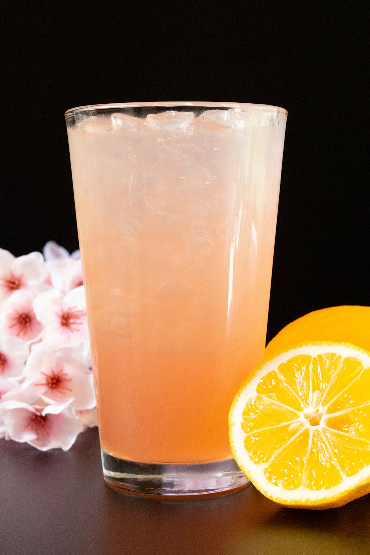 A pint glass filled with a light pink lemonade. Cherry blossoms and a sliced lemon are out of focus in the background.