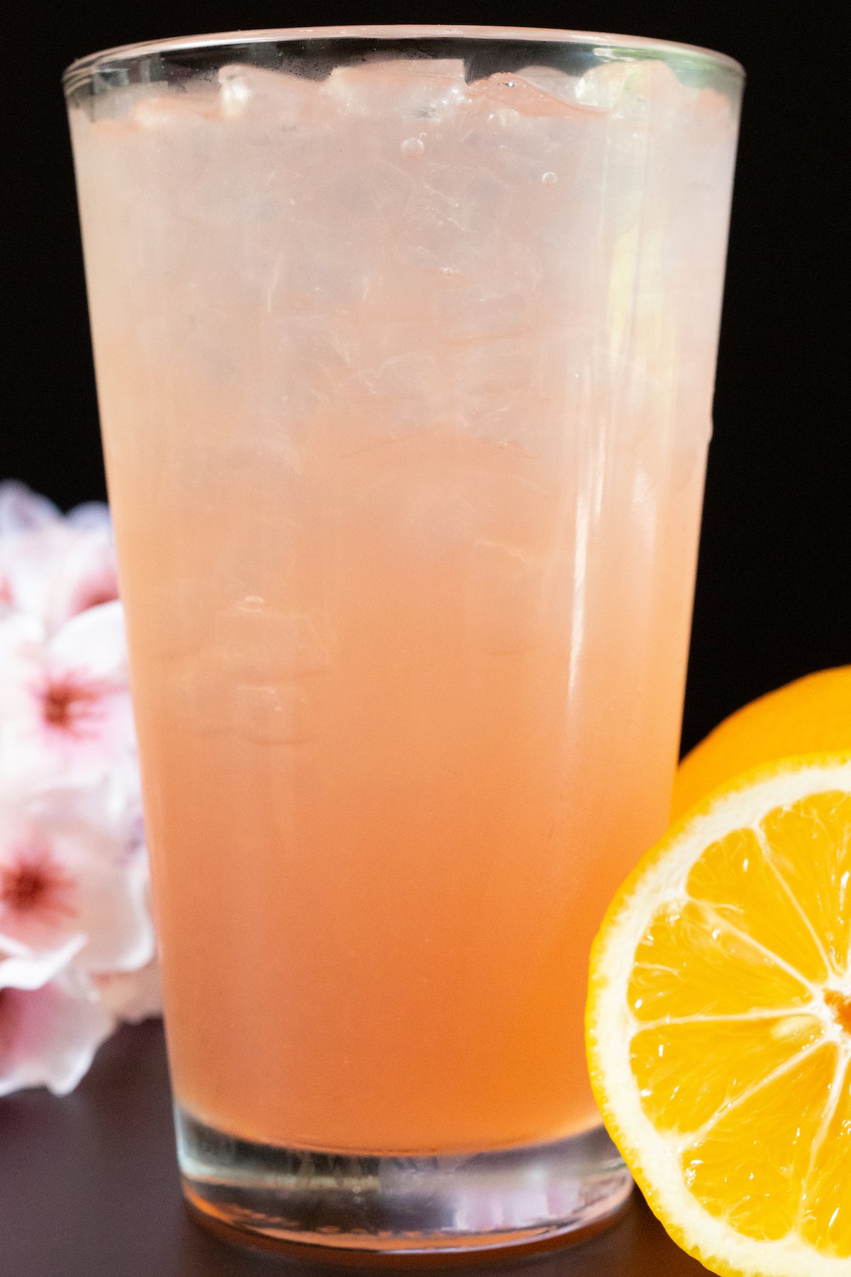 A frosty pint glass is filled with a light pink sakura flavored lemonade. A sliced lemon is in focus on the right, and cherry blossoms out of focus in the background on the left.