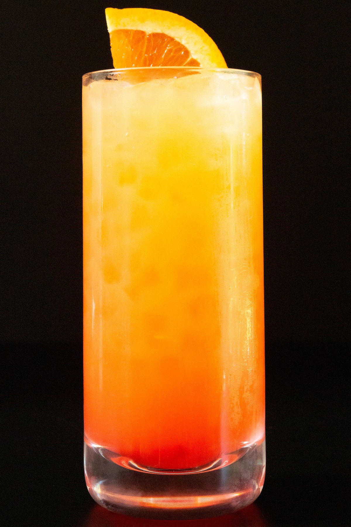 A high ball glass is filled with orange juice and grenadine that makes it look like a sunrise. A sliced orange is used as garnish.